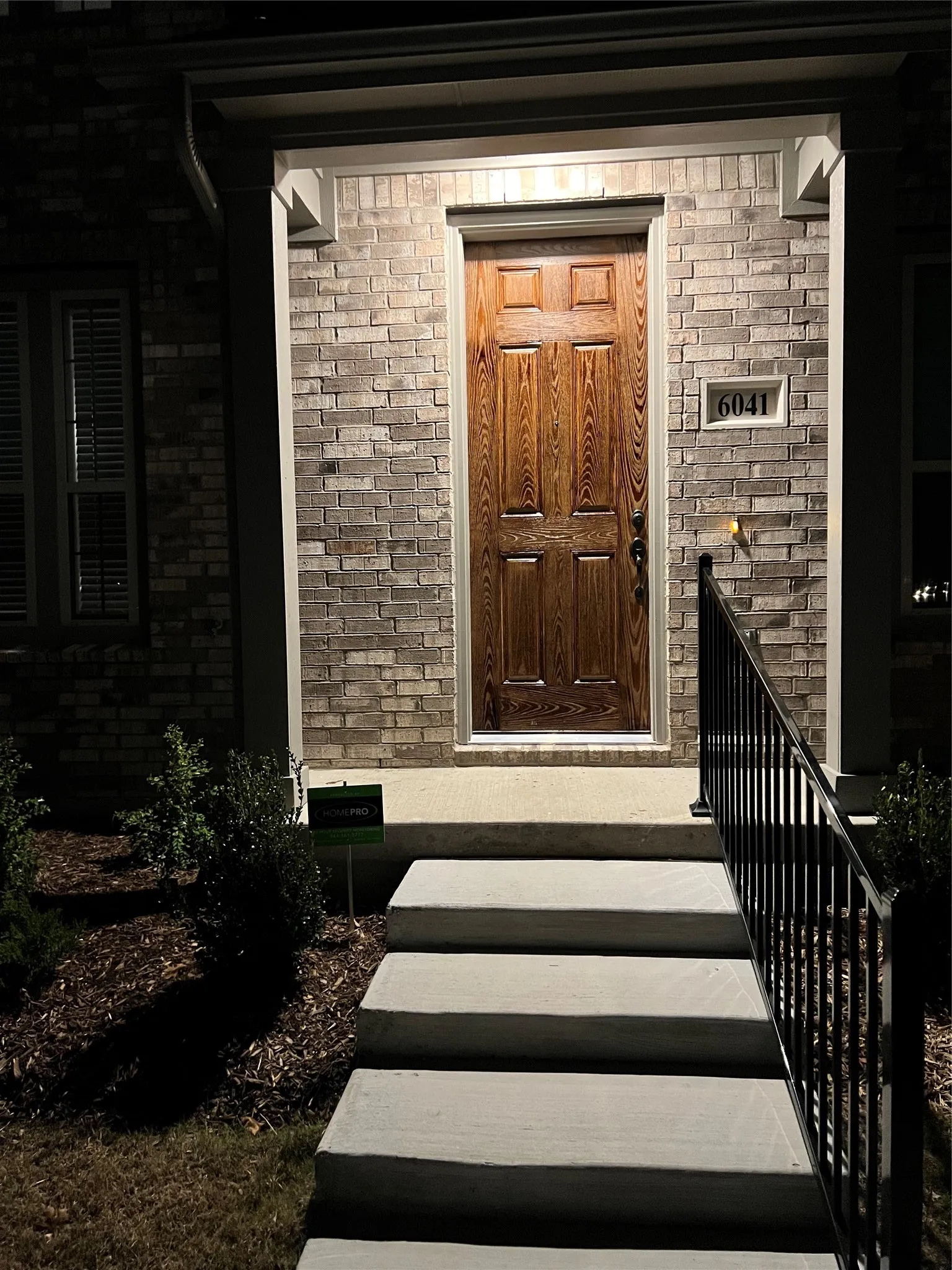 Entryway featuring a paneled wood-finish door, brick facade, concrete steps, wrought iron railing, and manicured landscaping