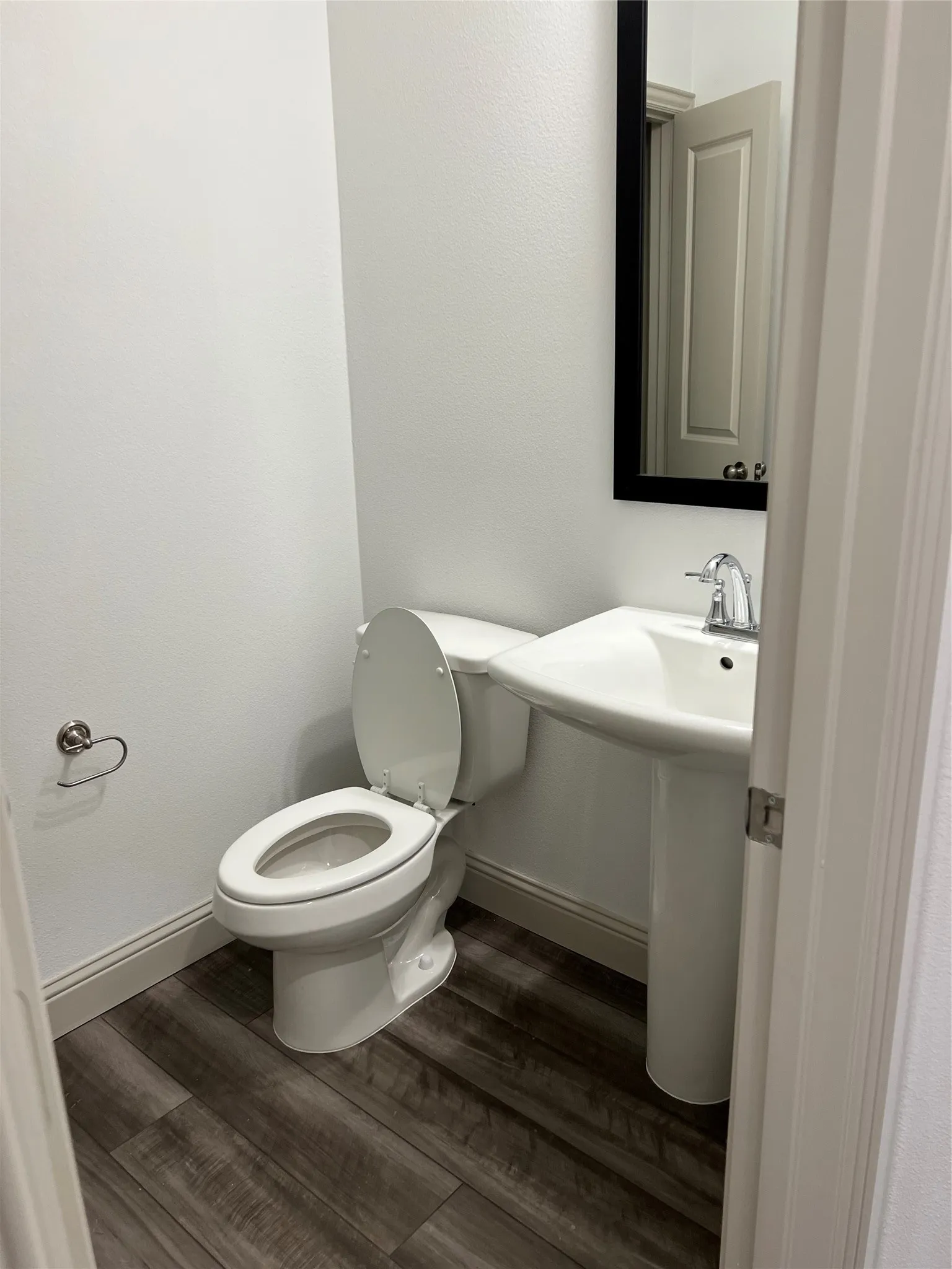 Compact powder room featuring a pedestal sink with a chrome faucet, a white toilet, wood-finish flooring, and a framed mirror
