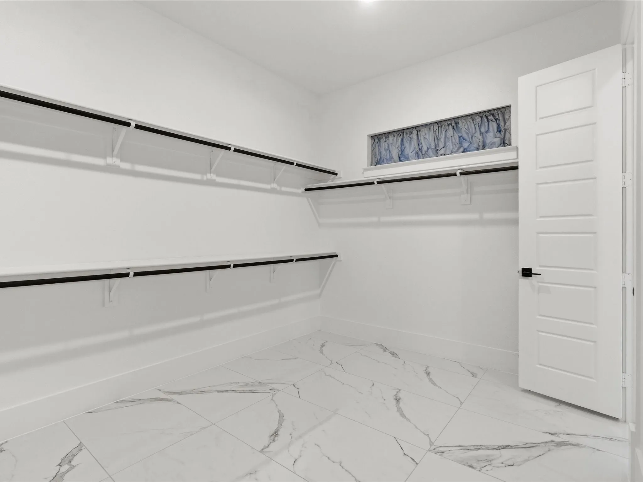 Spacious walk-in closet featuring white marble-look tile flooring, extensive built-in shelving, hanging rods, and a high horizontal window