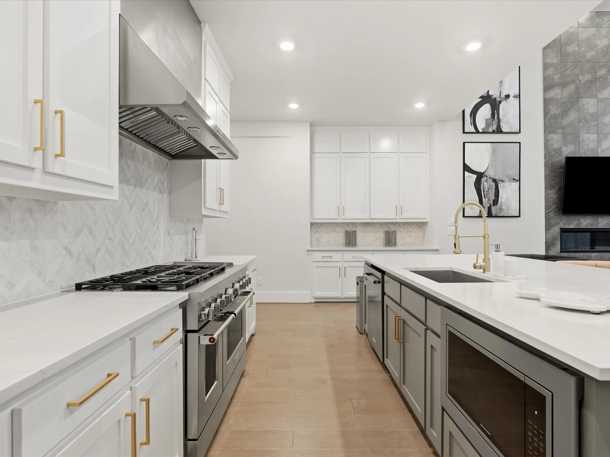 Contemporary kitchen featuring white cabinetry with gold-tone hardware, a stainless steel range with matching hood, a white herringbone tile backsplash, and a large central island with a built-in microwave and sink