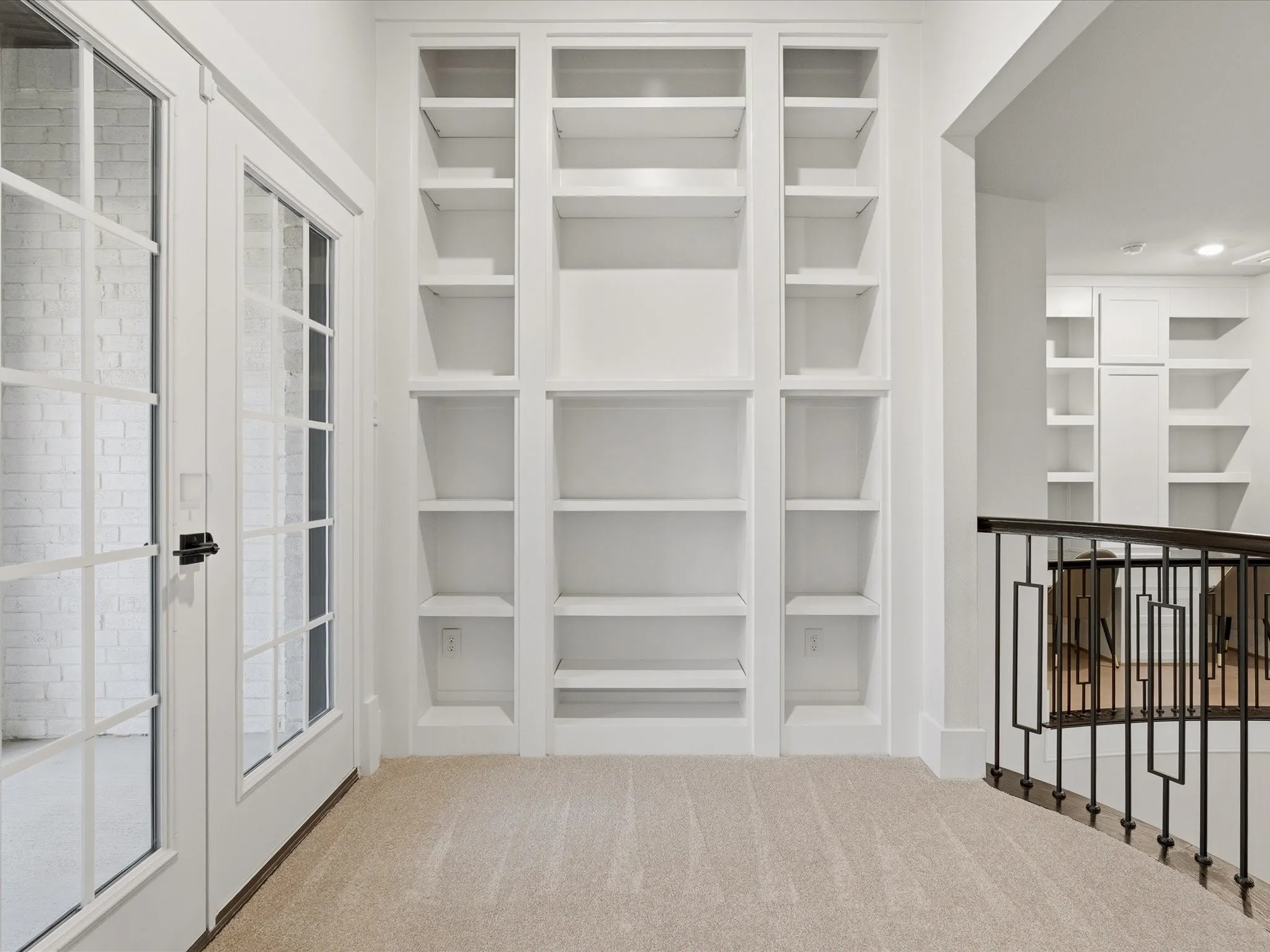 Built-in shelving with multiple compartments, French doors with grid-patterned glass, light-colored carpeting, and a wrought iron railing with geometric designs