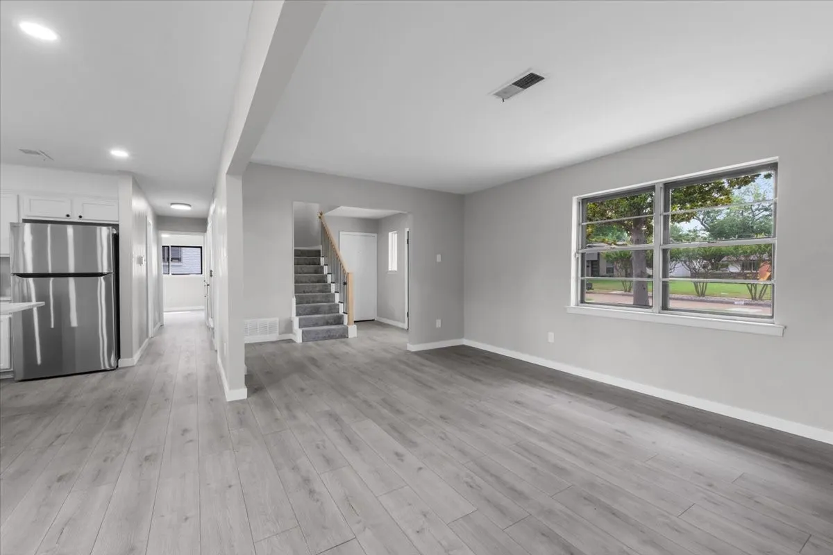 Open-concept living area featuring wood-finish flooring, recessed lighting, a large window, a staircase with carpeted treads, and a partial view of the kitchen with white cabinetry and a stainless steel refrigerator