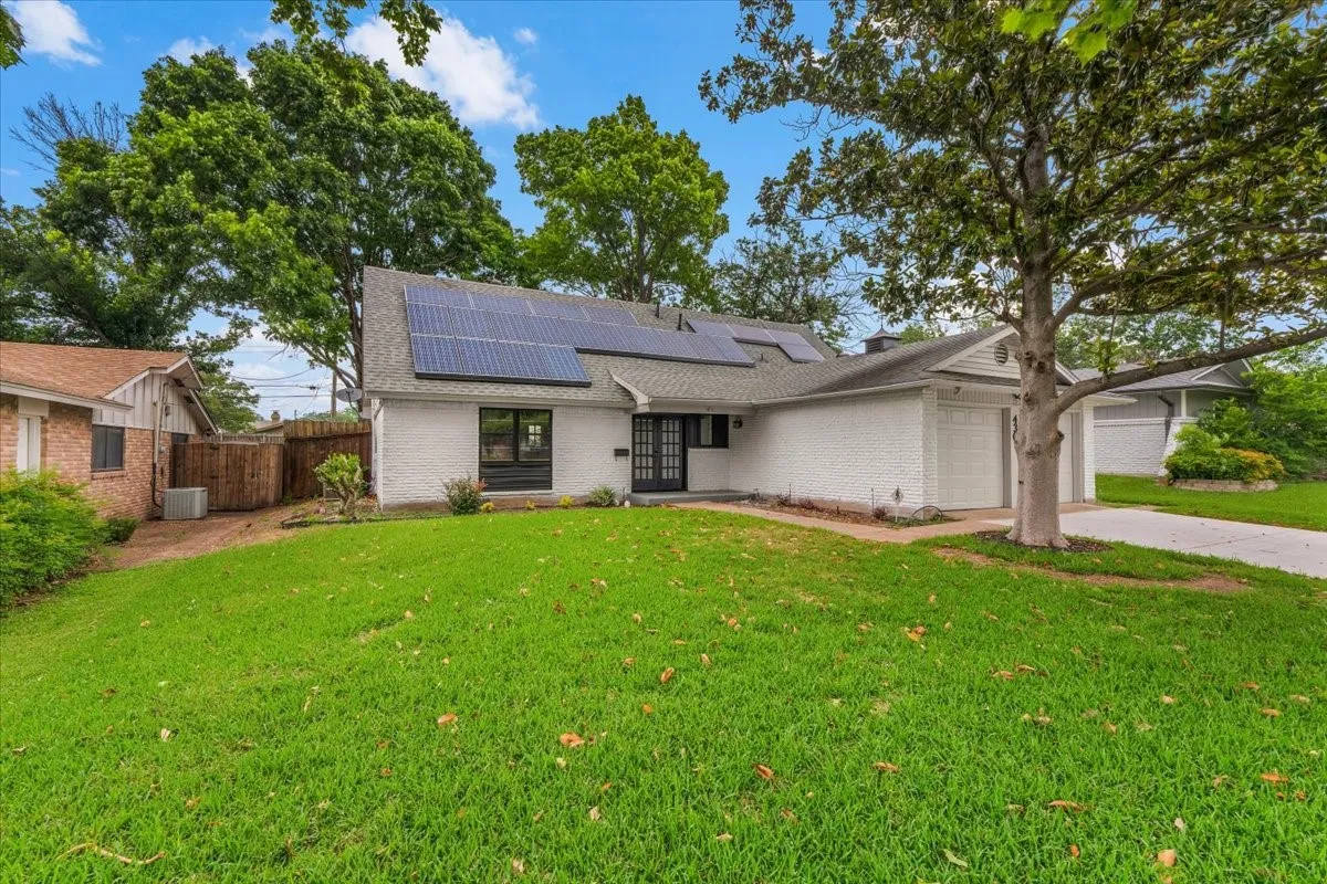 White painted brick exterior featuring a front entry with a black security door, an attached garage, solar panels on the roof, and a mature front tree
