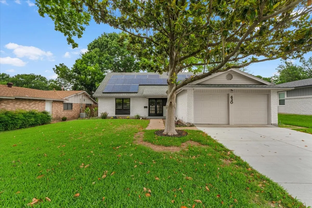 White brick exterior with a two-car garage, solar panels, mature front yard tree, and an entry with a dark door and side windows