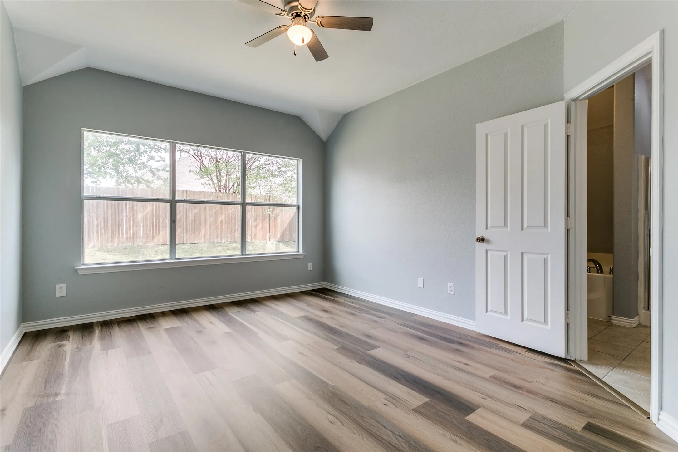 Vaulted ceiling room featuring a multi-pane window overlooking a wood fence, wood-finish flooring, and a ceiling fan