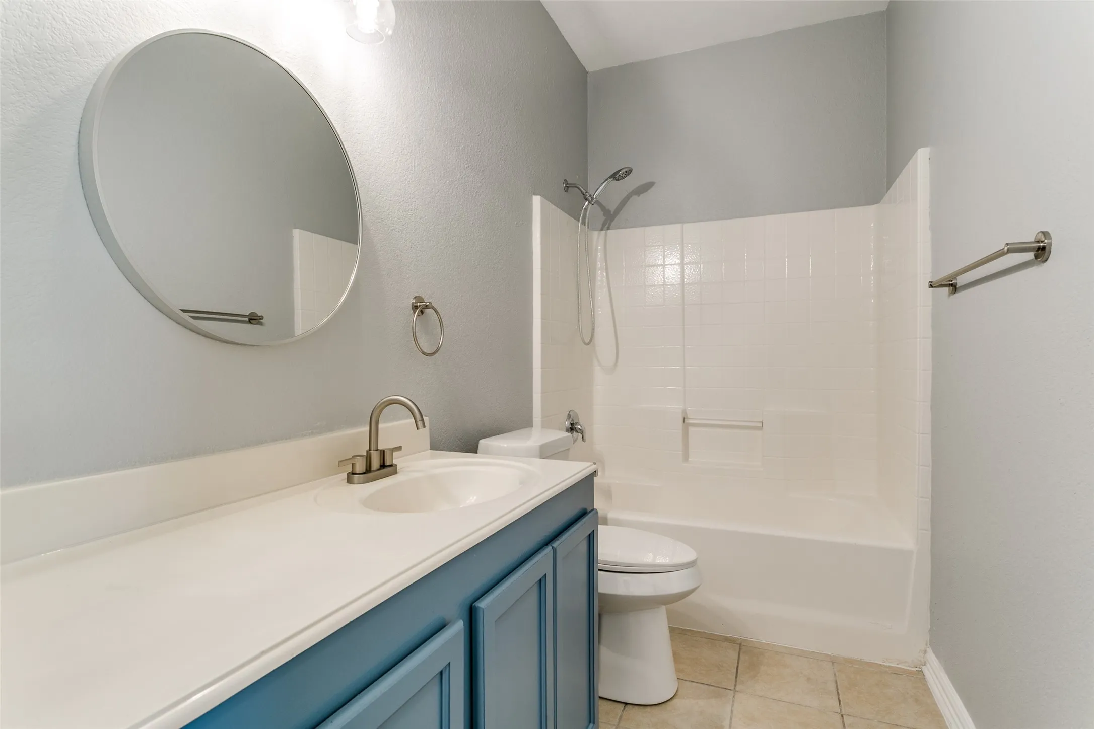 Bathroom featuring a blue vanity with an integrated white sink, a circular mirror, a shower-tub combination with white tile surround, and neutral tile flooring