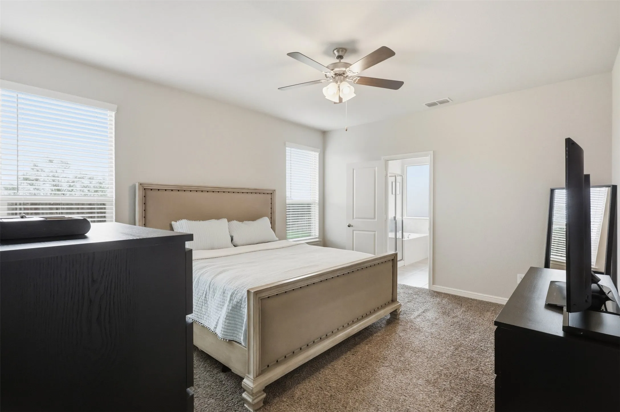 Carpeted bedroom with a ceiling fan, two windows with blinds, and a doorway opening to a bathroom featuring a bathtub