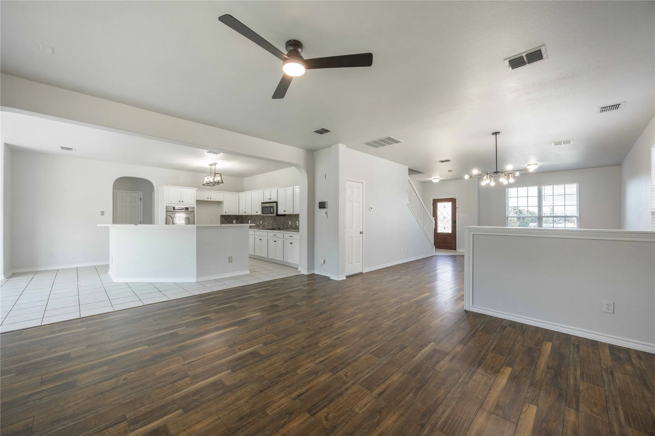 Expansive living area with wood-finish flooring, featuring a contemporary ceiling fan, an elegant multi-light chandelier, and a partial wall separating spaces