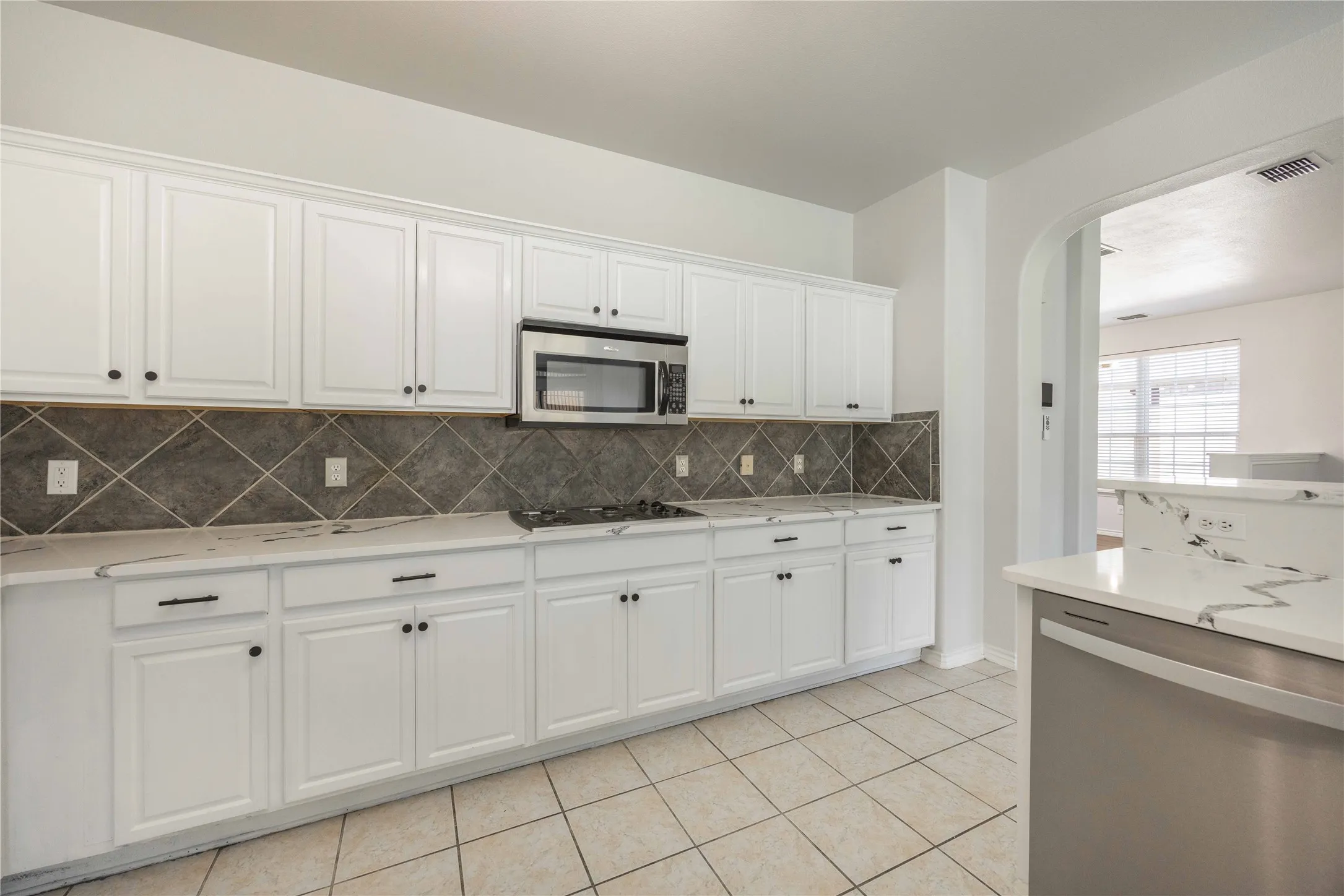 Kitchen featuring white cabinetry with dark hardware, a stainless steel microwave, a gas cooktop, patterned tile backsplash, and light-toned tile flooring