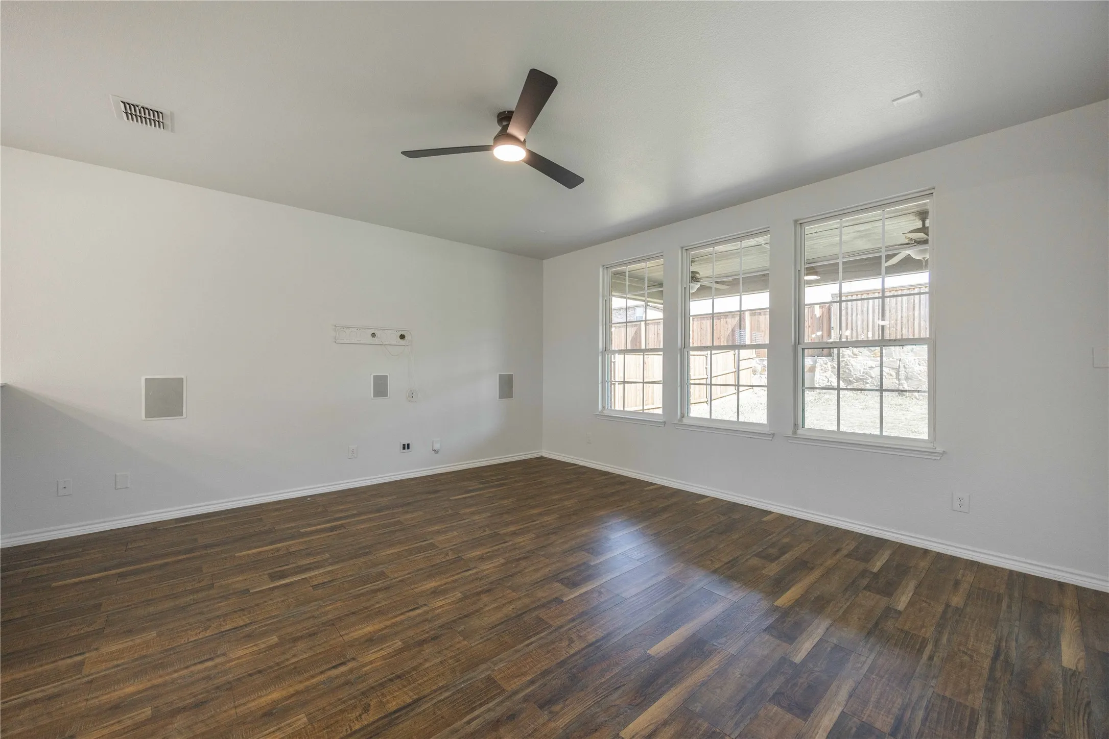 Spacious room featuring wood-finish flooring, three large windows, a ceiling fan with integrated lighting, and built-in wall speakers