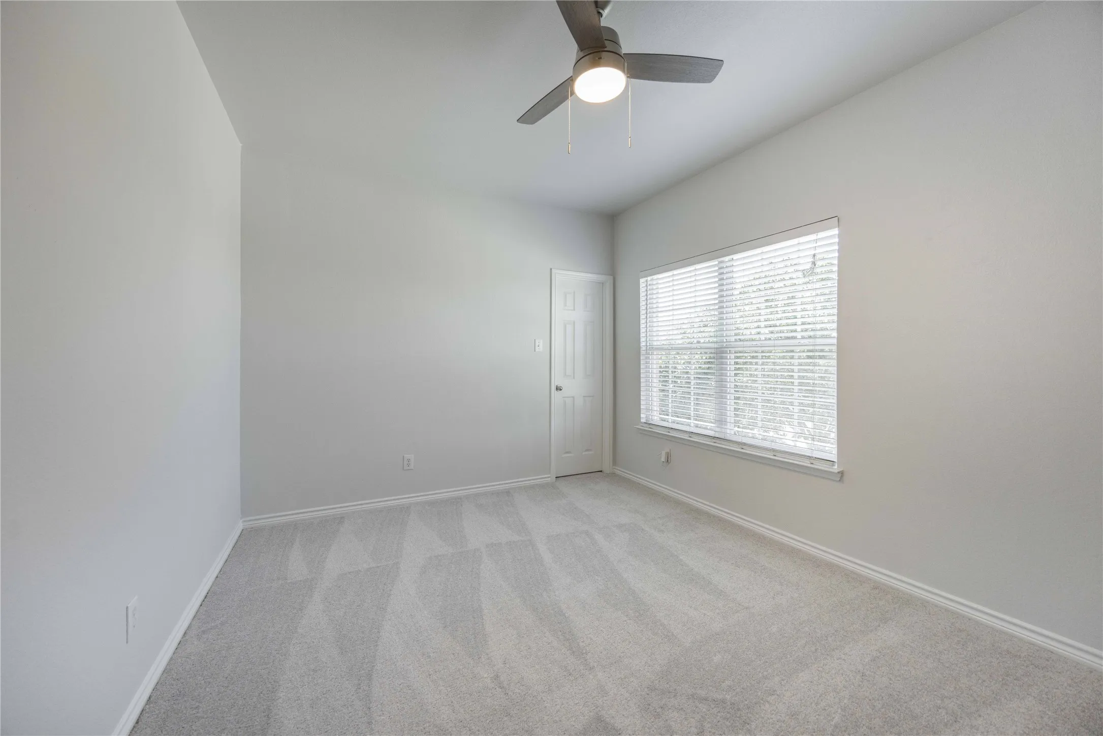 Spacious room featuring light gray carpeting, a large window with blinds, and a ceiling fan