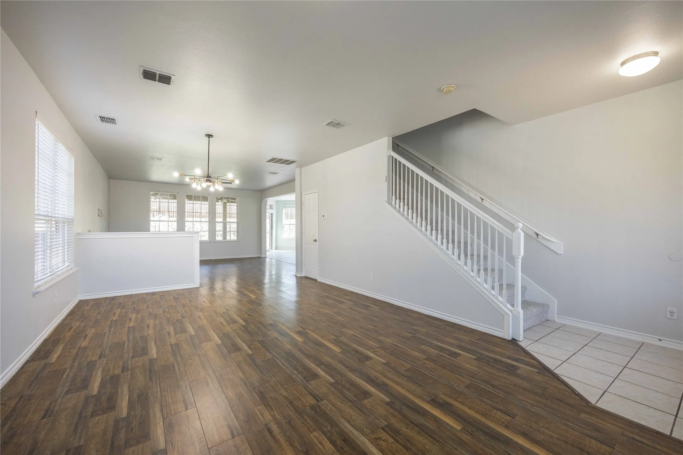 Spacious open-concept living area featuring wood-finish flooring, multiple windows with blinds, a prominent staircase with white railings and carpeted treads, a contemporary multi-bulb chandelier, and neutral wall paint