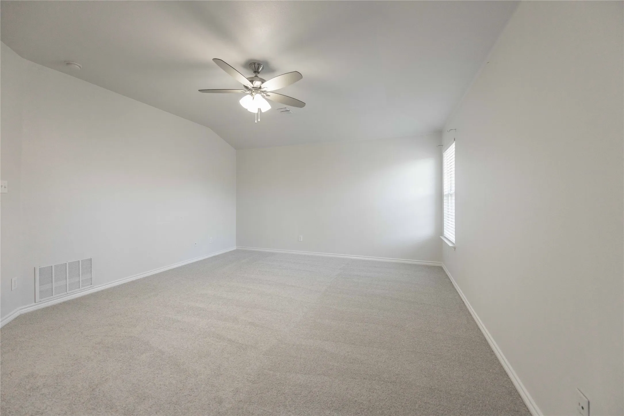 Spacious room featuring light gray carpeting, neutral wall finishes, a ceiling fan with integrated lighting, a single window with blinds, and white baseboards