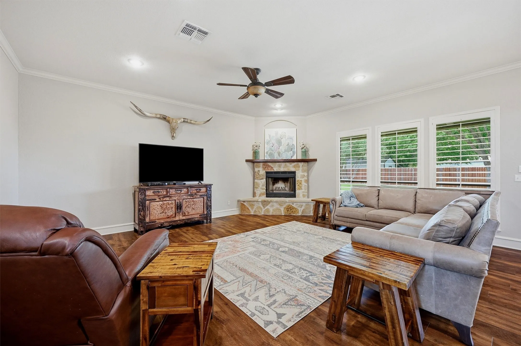 Spacious living area featuring wood-finish flooring, a corner stone fireplace with a wood mantle, and three large windows with plantation shutters
