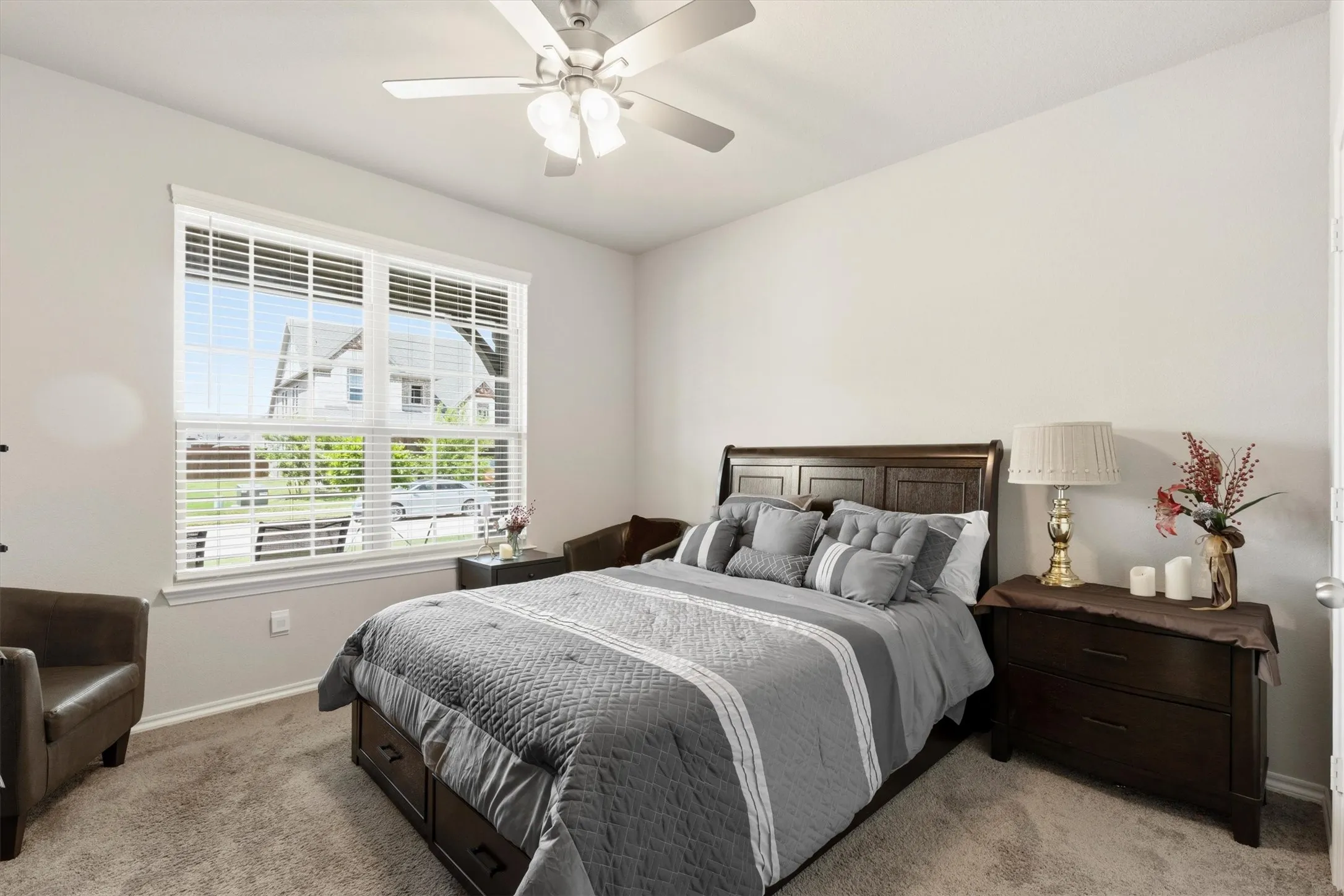 Bright bedroom featuring a large window with white blinds, neutral wall tones, textured carpeting, and a ceiling fan with integrated lighting