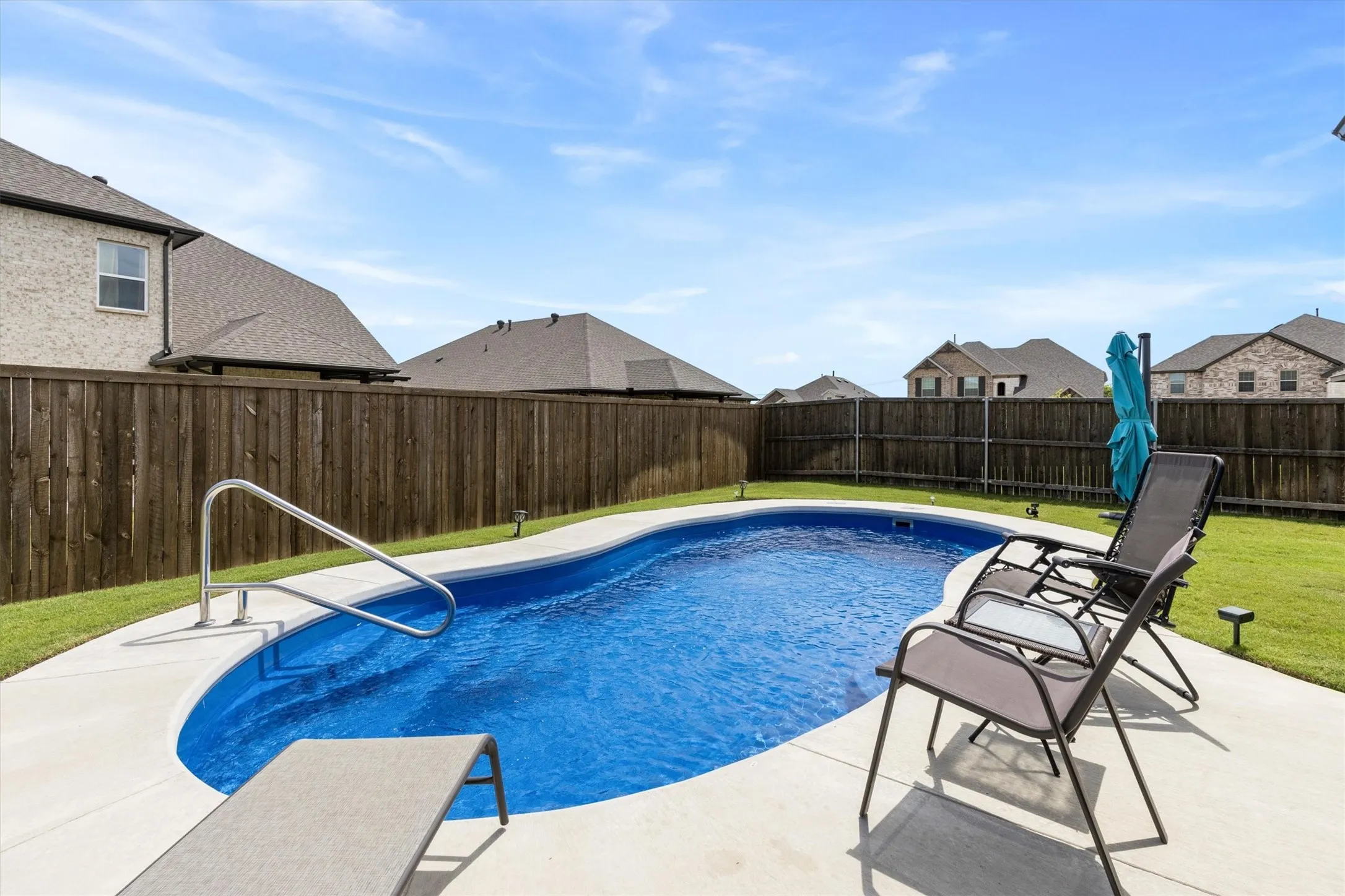 Private backyard featuring a curved inground swimming pool with a concrete surround, metal handrail, and perimeter landscape lighting