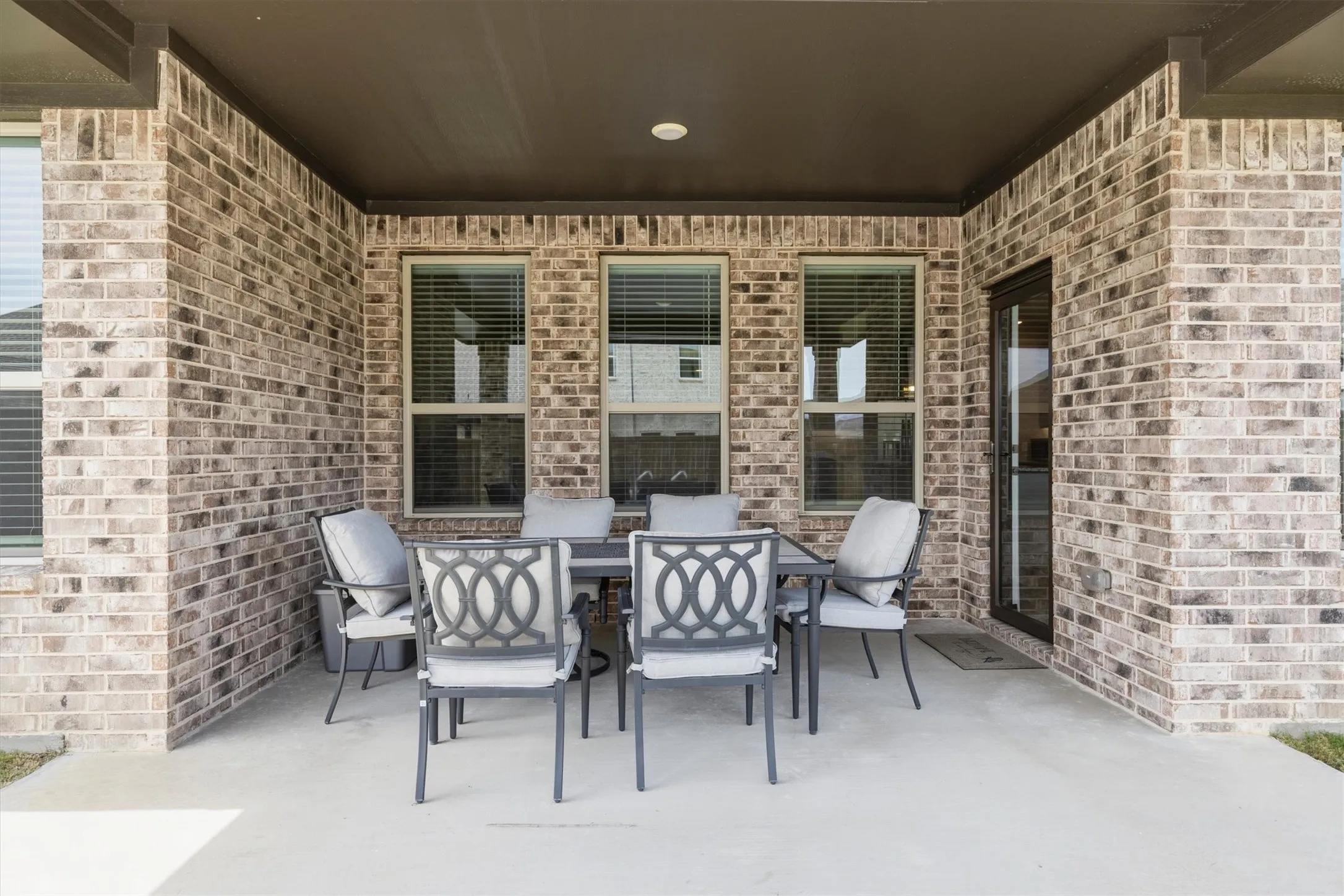 Covered concrete patio featuring a brick exterior, multiple windows with blinds, a recessed light, and an exterior door with a dark frame