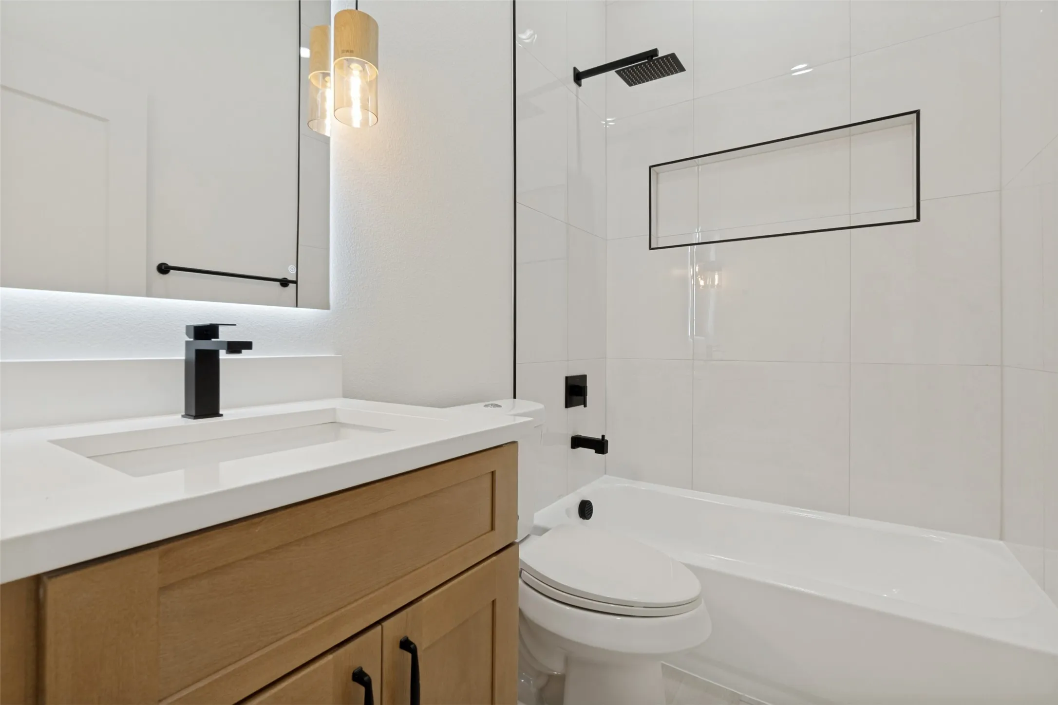 Contemporary bathroom featuring a light wood-finish vanity with an integrated sink, matte black fixtures, an illuminated mirror, and a tiled shower-tub combination with a recessed niche