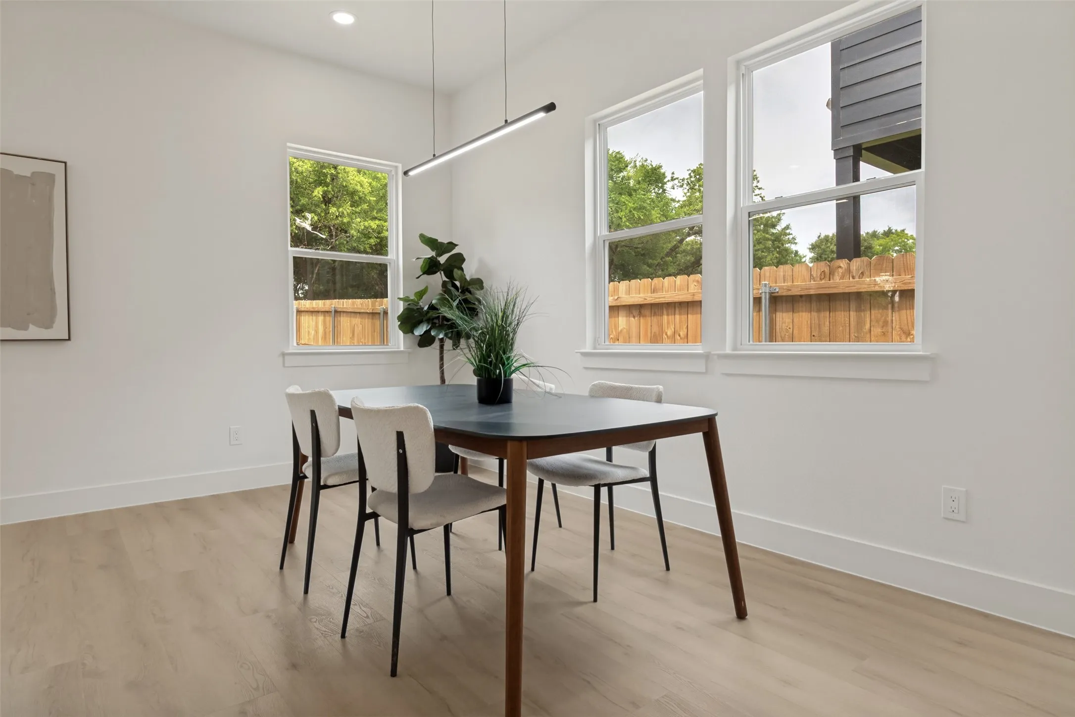 Dining area featuring a linear pendant light fixture, recessed ceiling light, three windows with white trim, light wood-finish flooring, and white baseboards