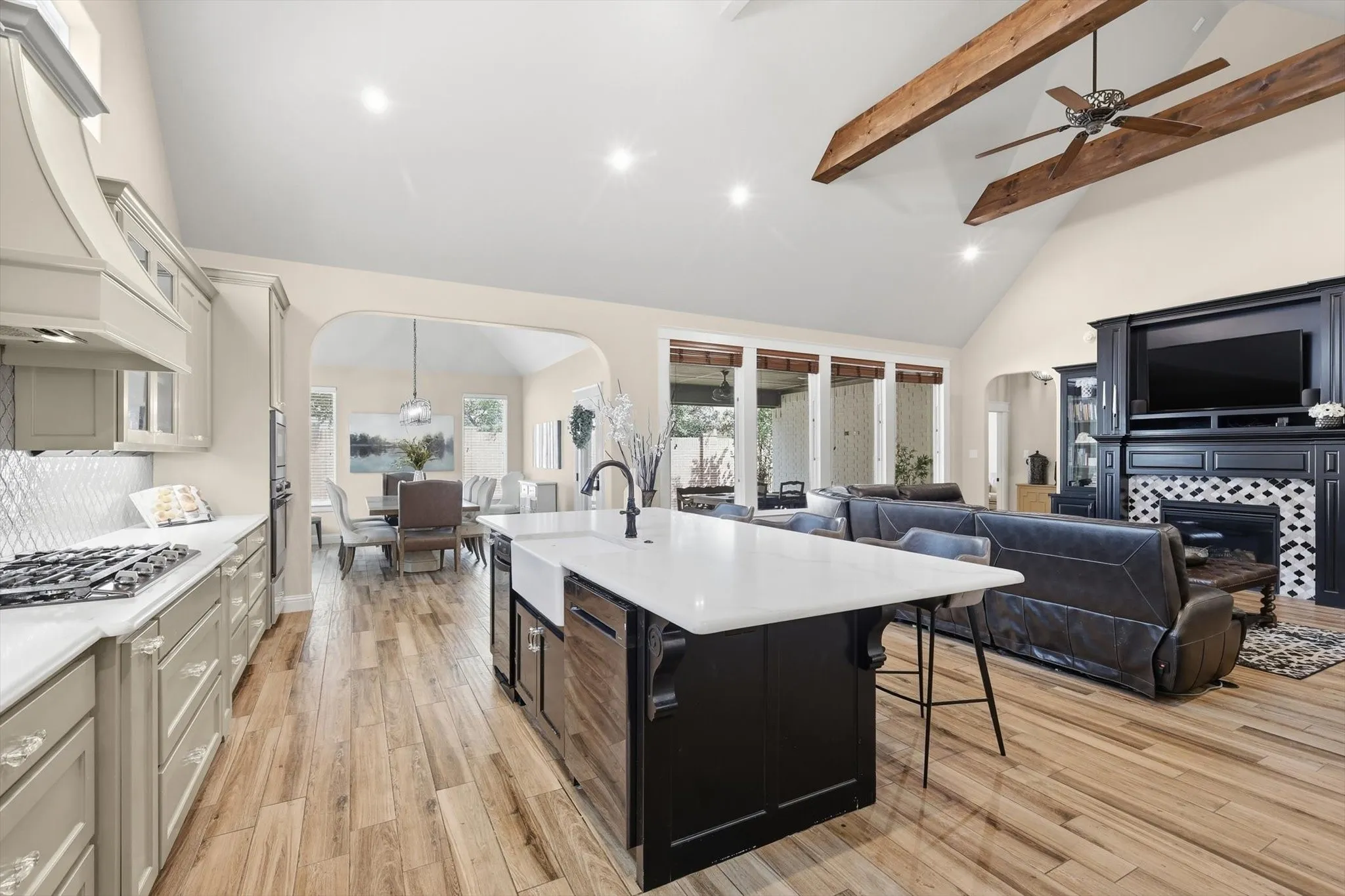 Open-concept living space featuring wood-finish flooring, a kitchen island with a farmhouse sink, a gas range with a tile backsplash, a fireplace with decorative tile, and exposed ceiling beams
