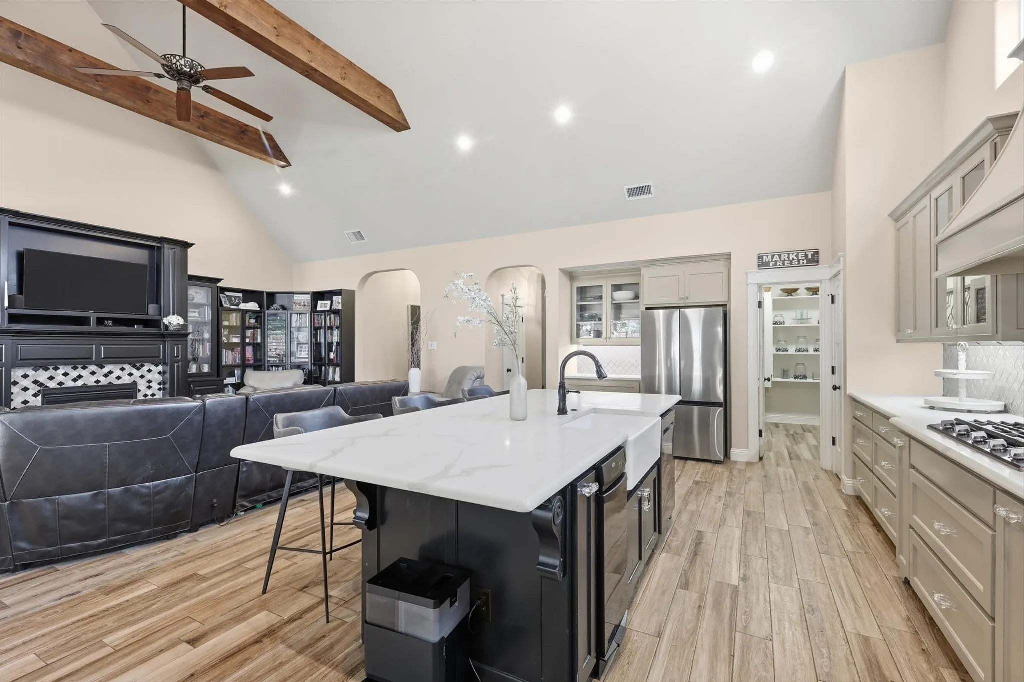 Open-concept living space featuring vaulted ceilings with exposed wood beams, wood-finish flooring, a kitchen island with a farmhouse sink and seating, stainless steel appliances, and a dedicated pantry