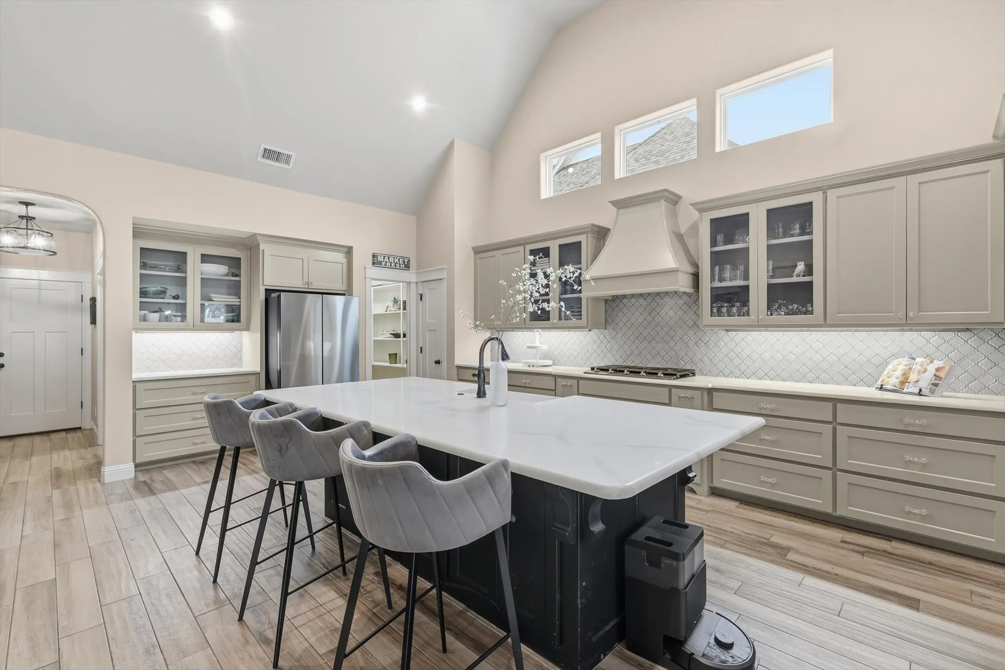 Spacious kitchen featuring a large central island with a white countertop, light wood-finish flooring, vaulted ceiling, and ample cabinetry with glass-front uppers