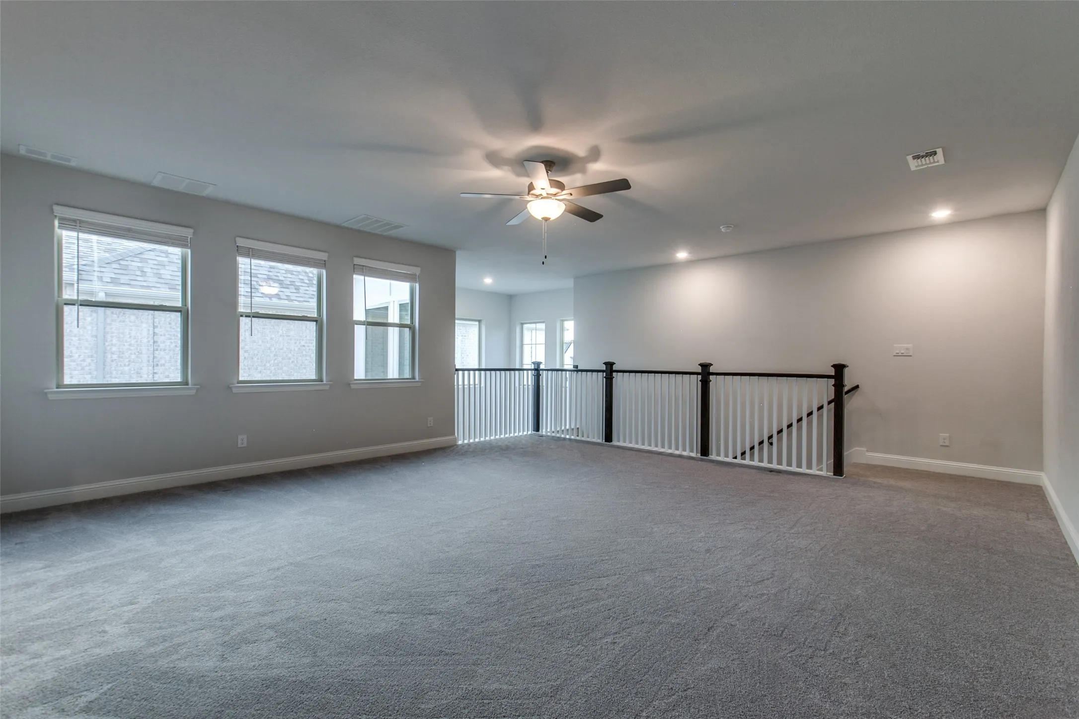 Spacious carpeted open-concept bonus room featuring three windows, a ceiling fan, recessed lighting, and a white and dark wood banister