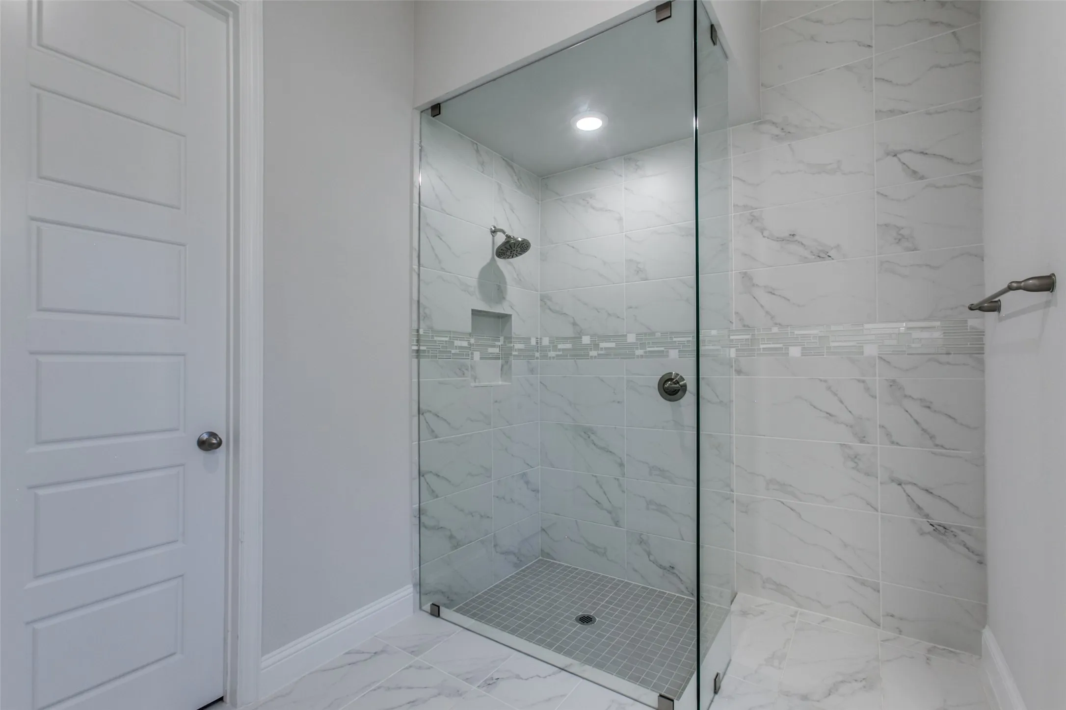 Spacious walk-in shower featuring clear glass enclosure, marble-look wall tiles with decorative glass accent band, and a tiled shower pan