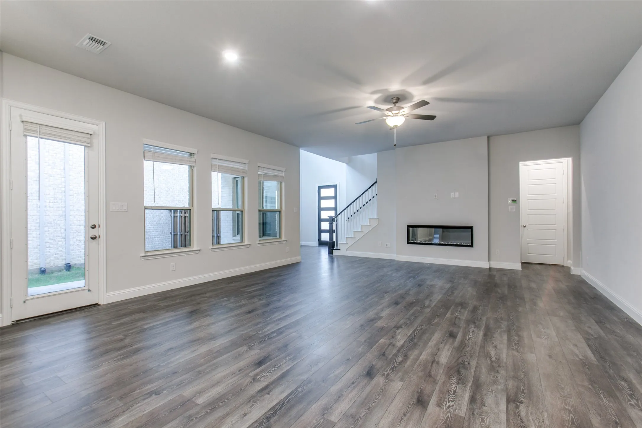 Spacious living area featuring wood-finish flooring, multiple windows, a ceiling fan with integrated lighting, a modern linear fireplace, and a staircase with open risers and dark handrails