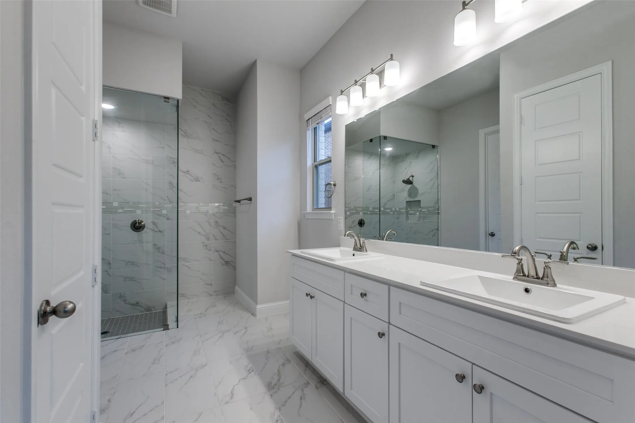Bathroom featuring a dual vanity with white cabinetry and integrated sinks, a large wall-mounted mirror, and a frameless glass shower with marble-look tile