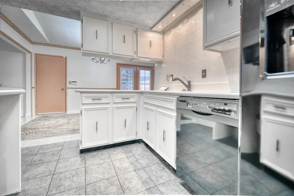 White cabinetry with silver hardware, light-toned countertop, stainless steel dishwasher, tile flooring, and a tiled backsplash