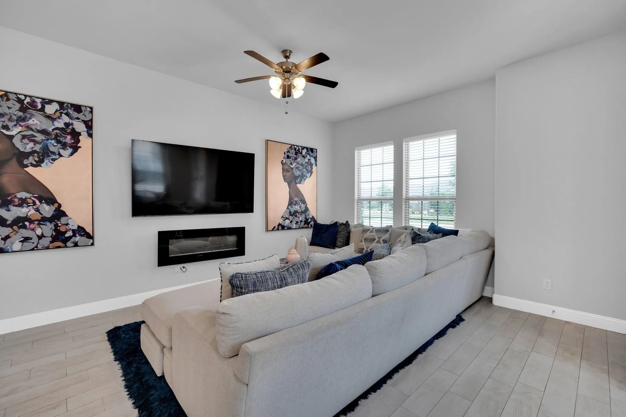 Bright living space featuring light gray walls, wood-finish flooring, two large windows with blinds, a wall-mounted fireplace, and a ceiling fan with integrated lighting