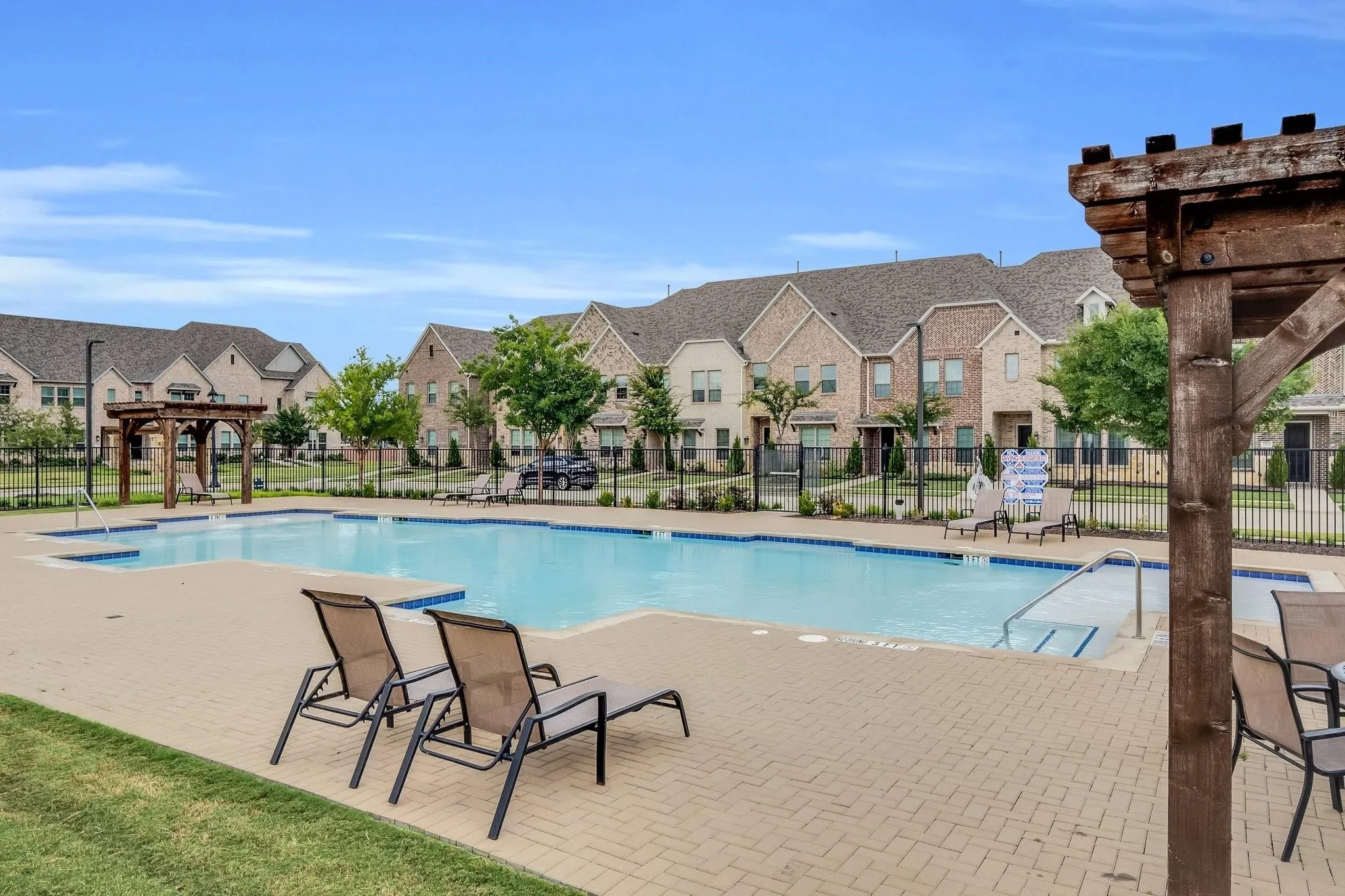 Community swimming pool with a paved deck, surrounded by townhomes featuring brick and siding exteriors, enclosed by a black metal fence, and accented by mature landscaping