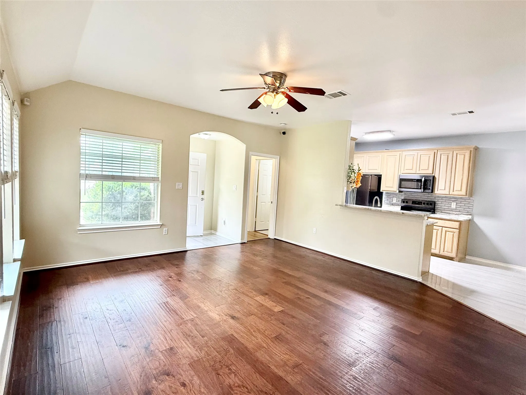 Open-concept living space with vaulted ceilings, rich wood-finish flooring, and a kitchen featuring light wood cabinetry, stainless steel appliances, and a tiled backsplash