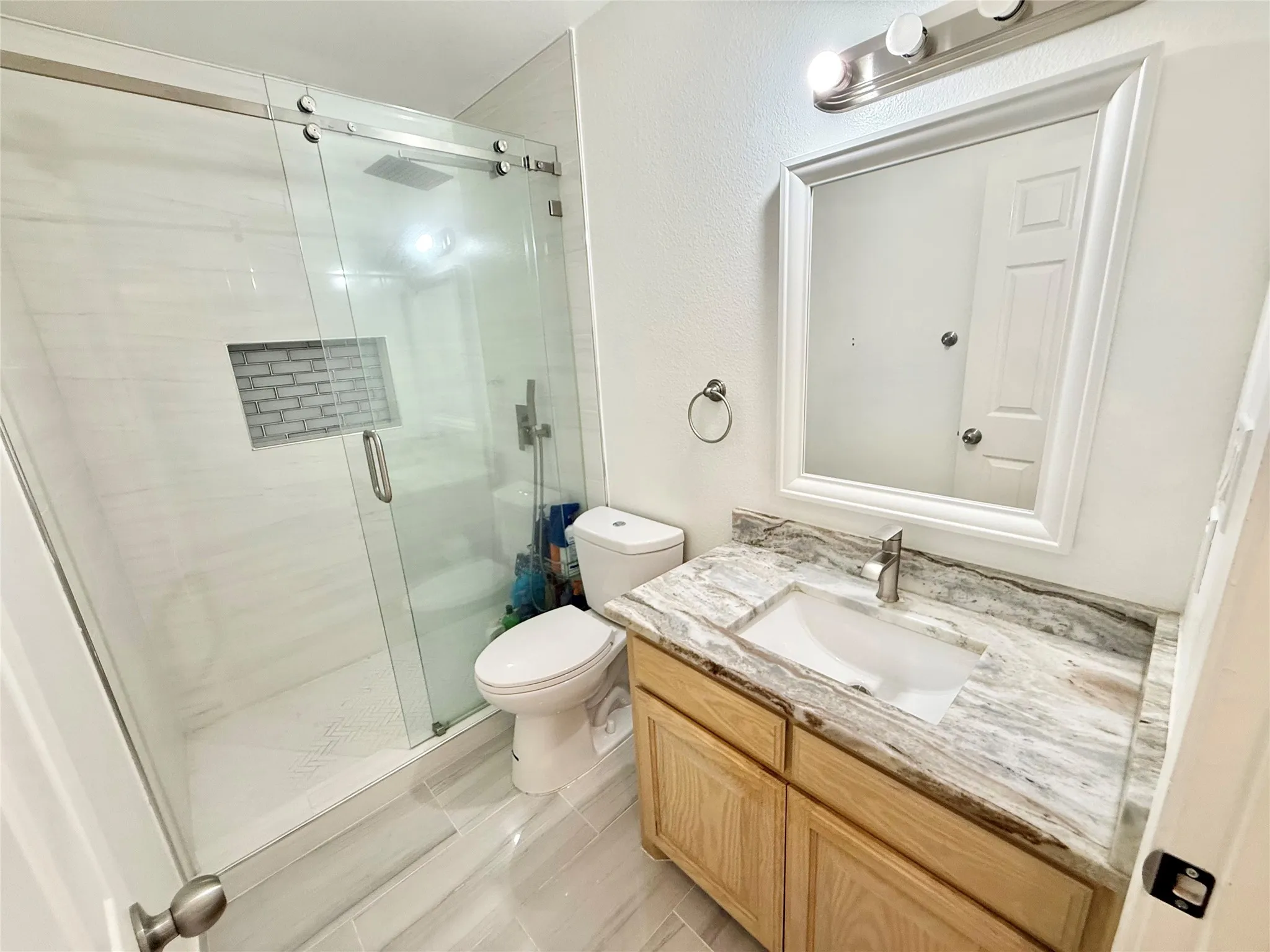 Contemporary bathroom featuring a glass-enclosed shower with a recessed tile niche, a wood-finish vanity with an integrated sink, a stone countertop, and light-toned tile flooring
