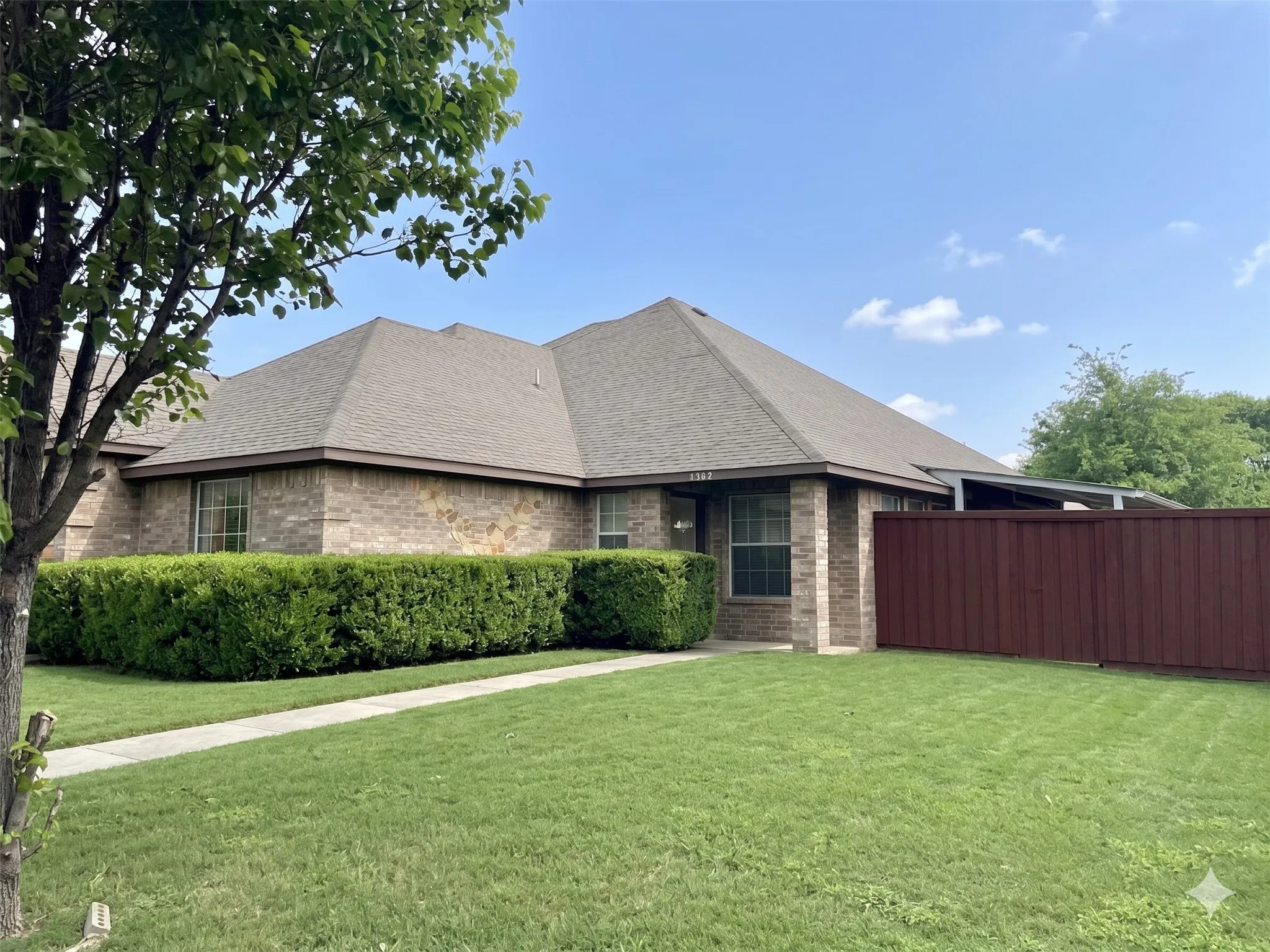 Brick exterior featuring a gray shingle roof, established landscaping, a concrete walkway, and a privacy fence