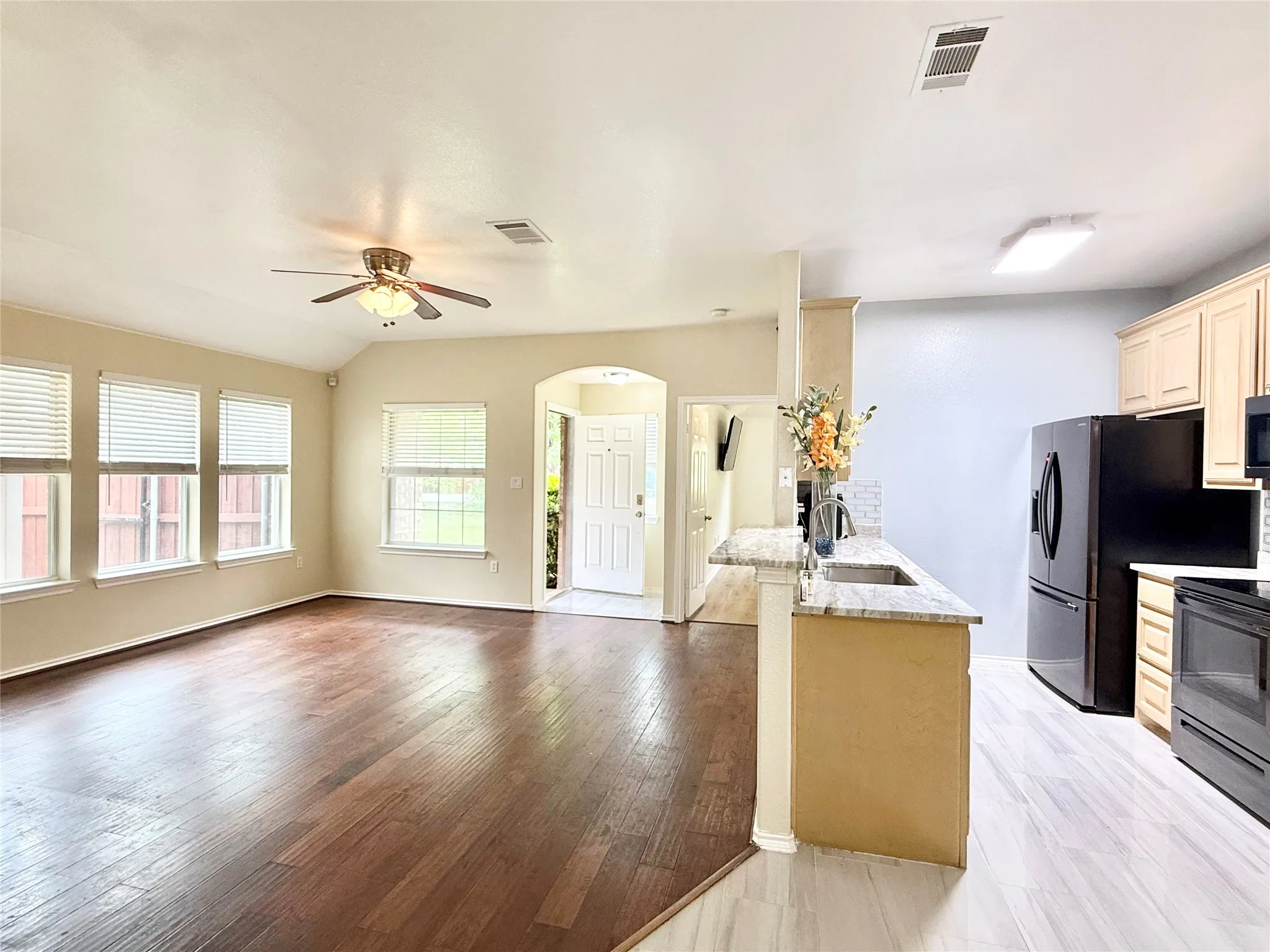 Open-concept living space featuring wood-finish flooring, multiple windows with blinds, a ceiling fan, and an integrated kitchen with a breakfast bar, light-toned cabinetry, and black appliances