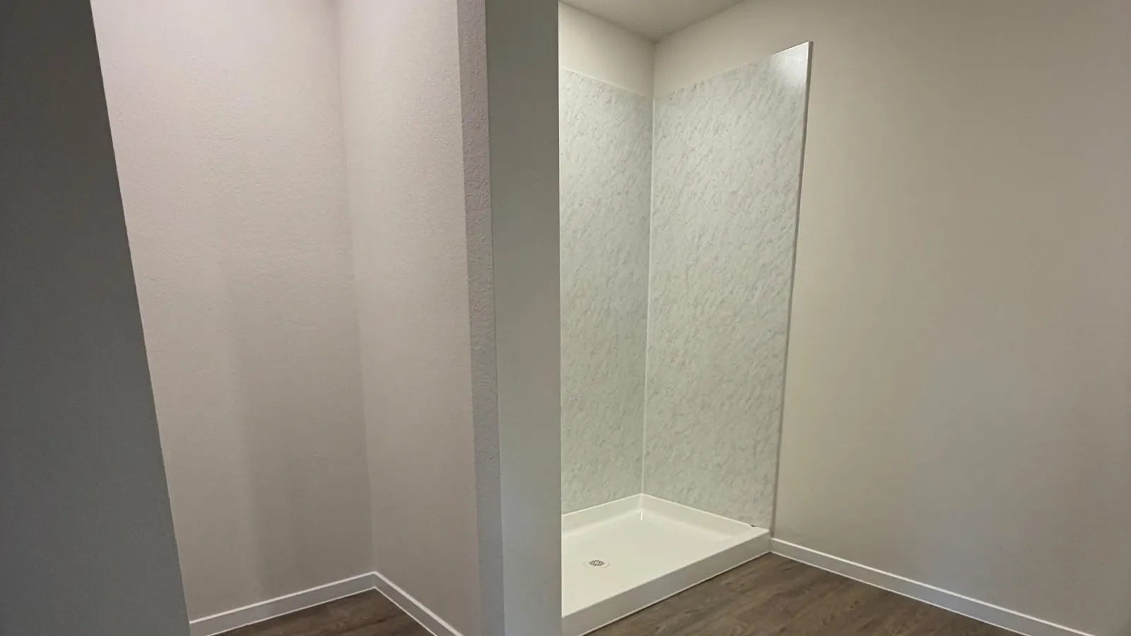 Shower with white base and textured wall panels, complemented by wood-finish flooring and white baseboards