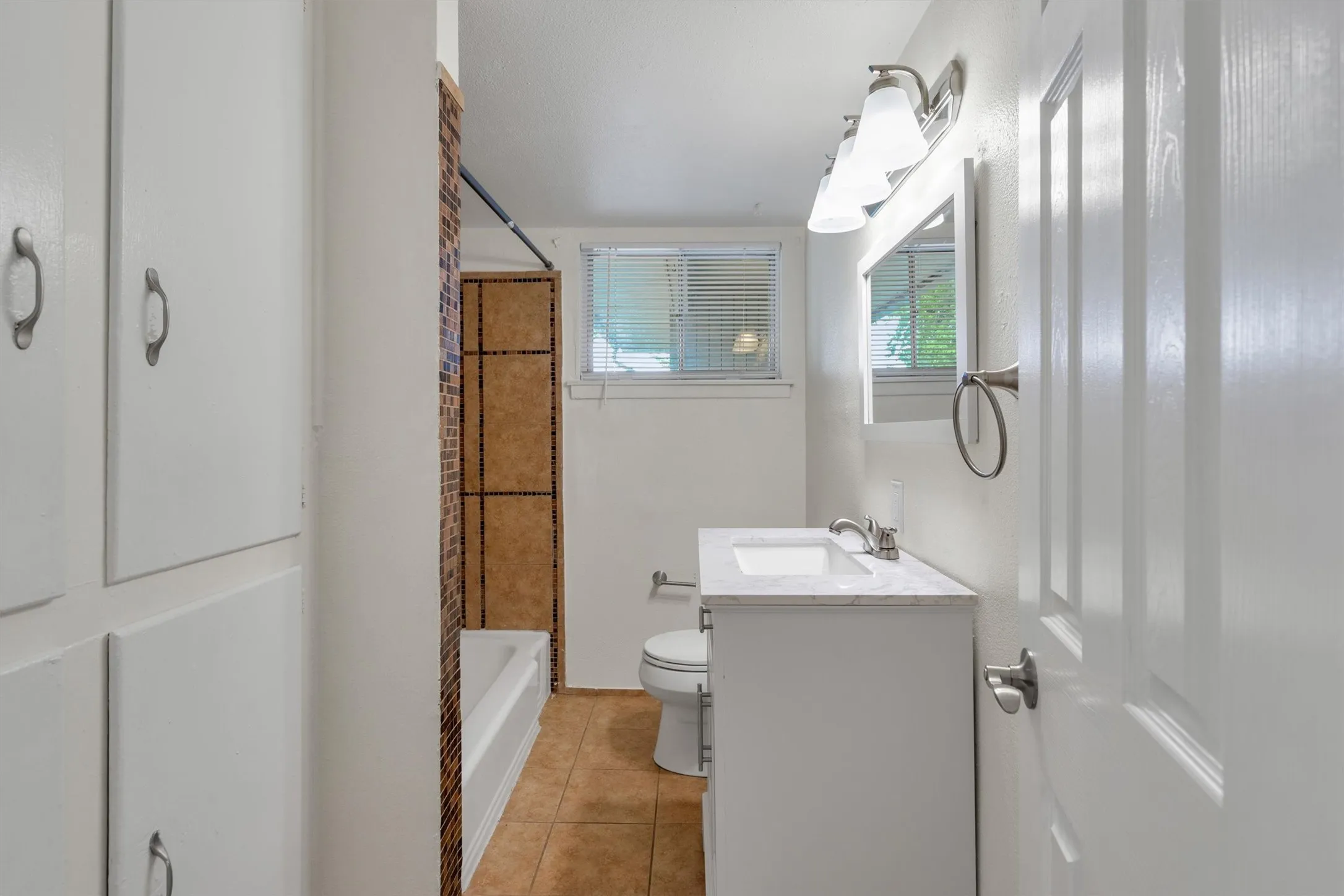 Bathroom featuring a white vanity with an integrated sink, a tiled tub surround, built-in white cabinetry, a window with blinds, and tile flooring