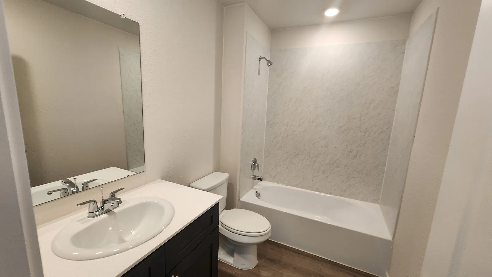 Bathroom featuring a white bathtub with a tile-patterned surround, a dark wood-finish vanity with a white countertop and integrated sink, a rectangular wall-mounted mirror, and wood-finish flooring