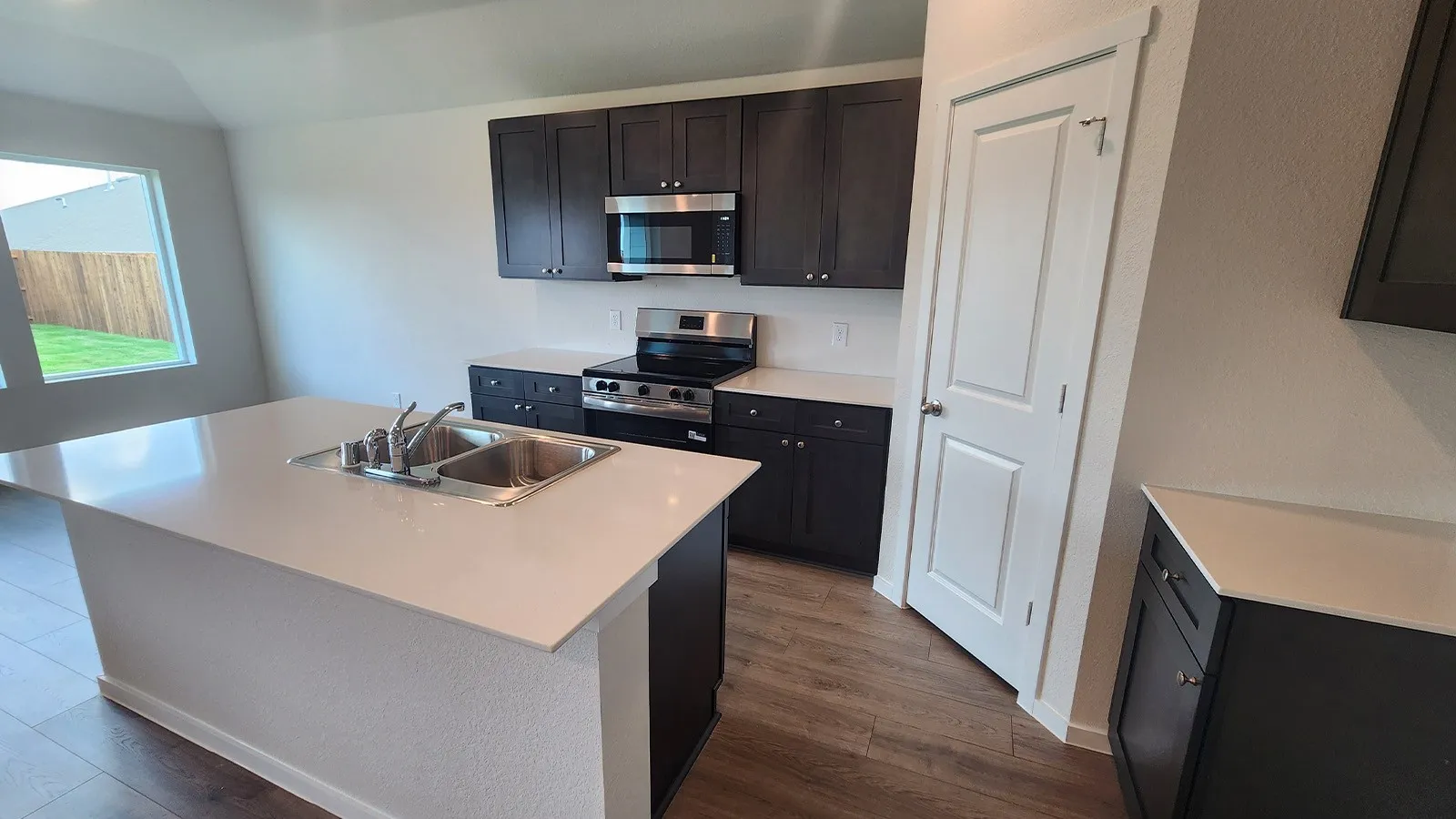 Kitchen featuring a central island with an undermount double basin stainless steel sink and white countertop, dark wood-finish cabinetry, stainless steel appliances, and wood-finish flooring