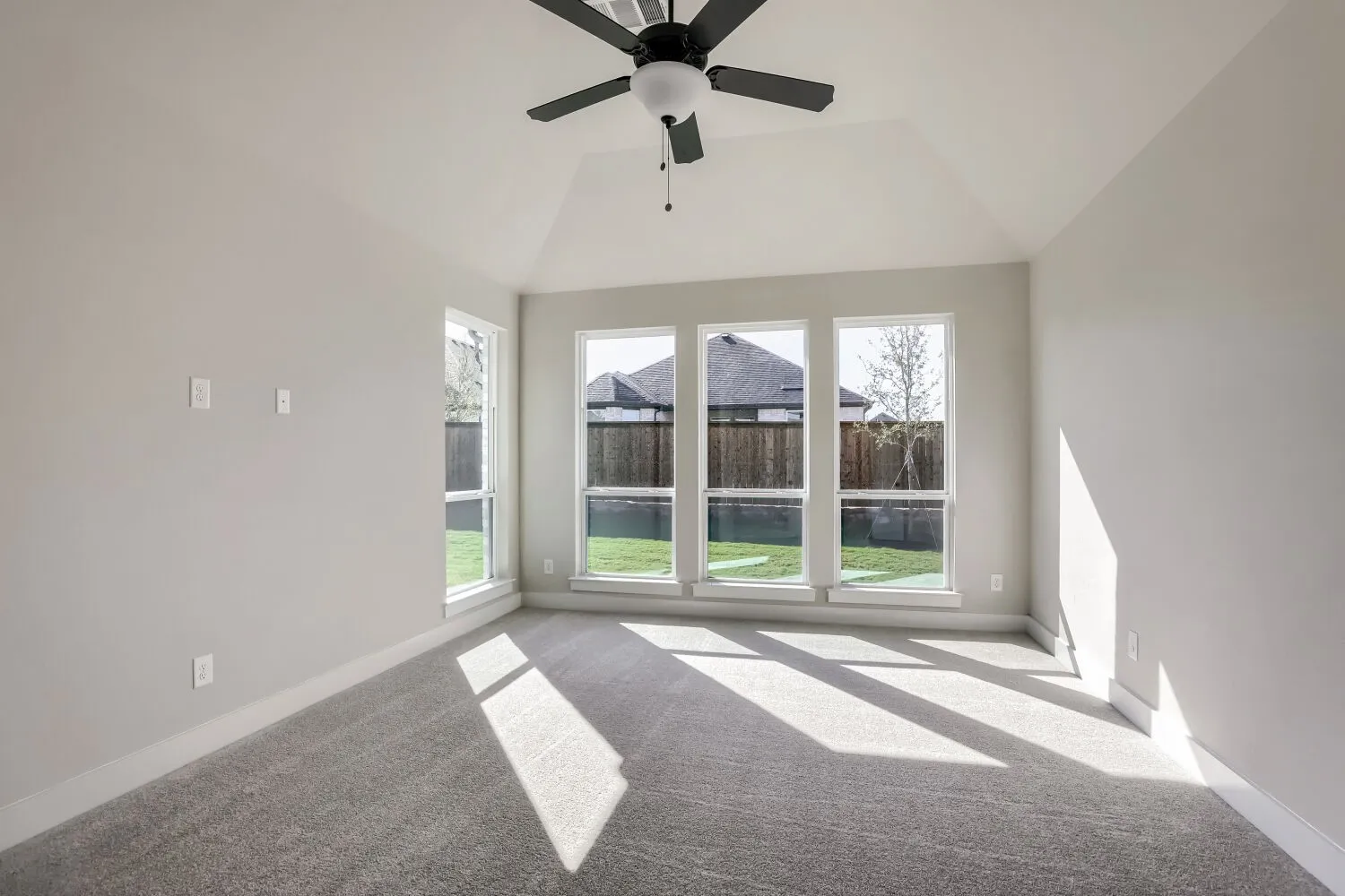 Vaulted ceiling with a ceiling fan, plush gray carpeting, and multiple windows overlooking a fenced backyard