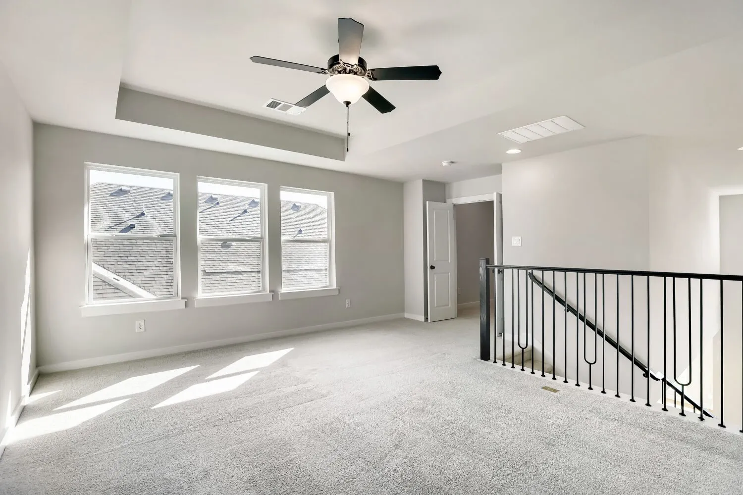 Carpeted open-concept space featuring three windows, a ceiling fan with light fixture, recessed lighting, and a black wrought iron railing