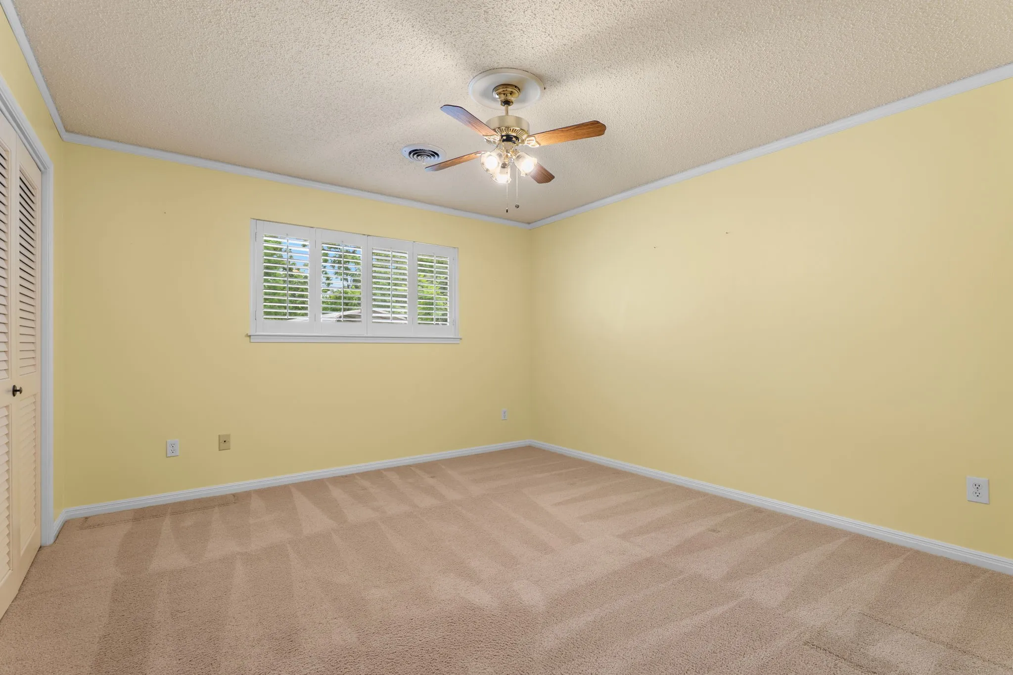 Carpeted interior space featuring a window with white louvered shutters, a ceiling fan with light fixture, and a built-in bifold closet