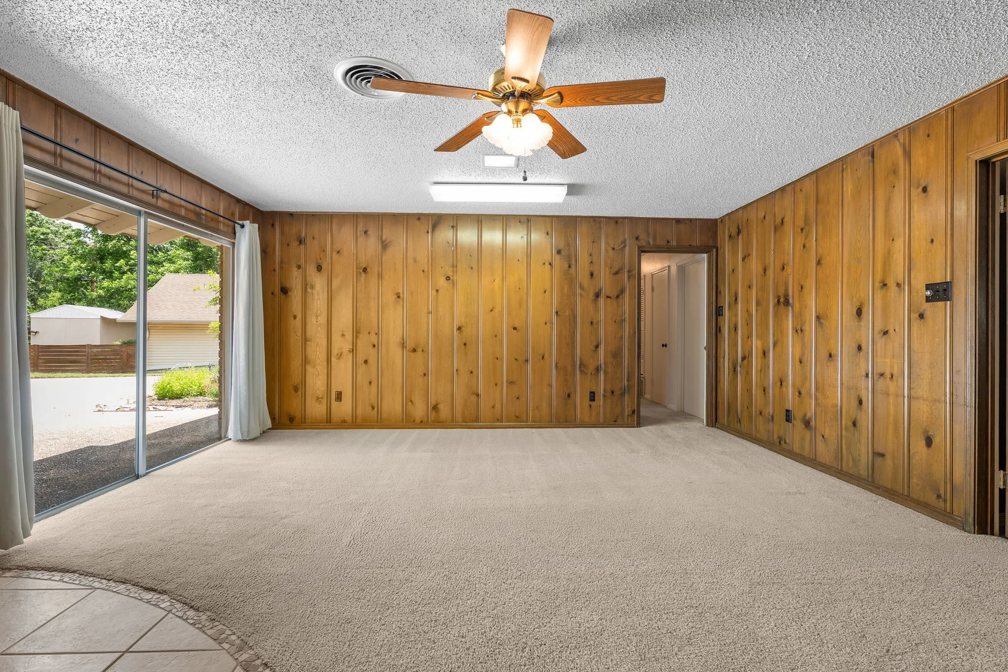Carpeted room featuring wood paneling, sliding glass doors, a ceiling fan with integrated lighting, and a textured ceiling