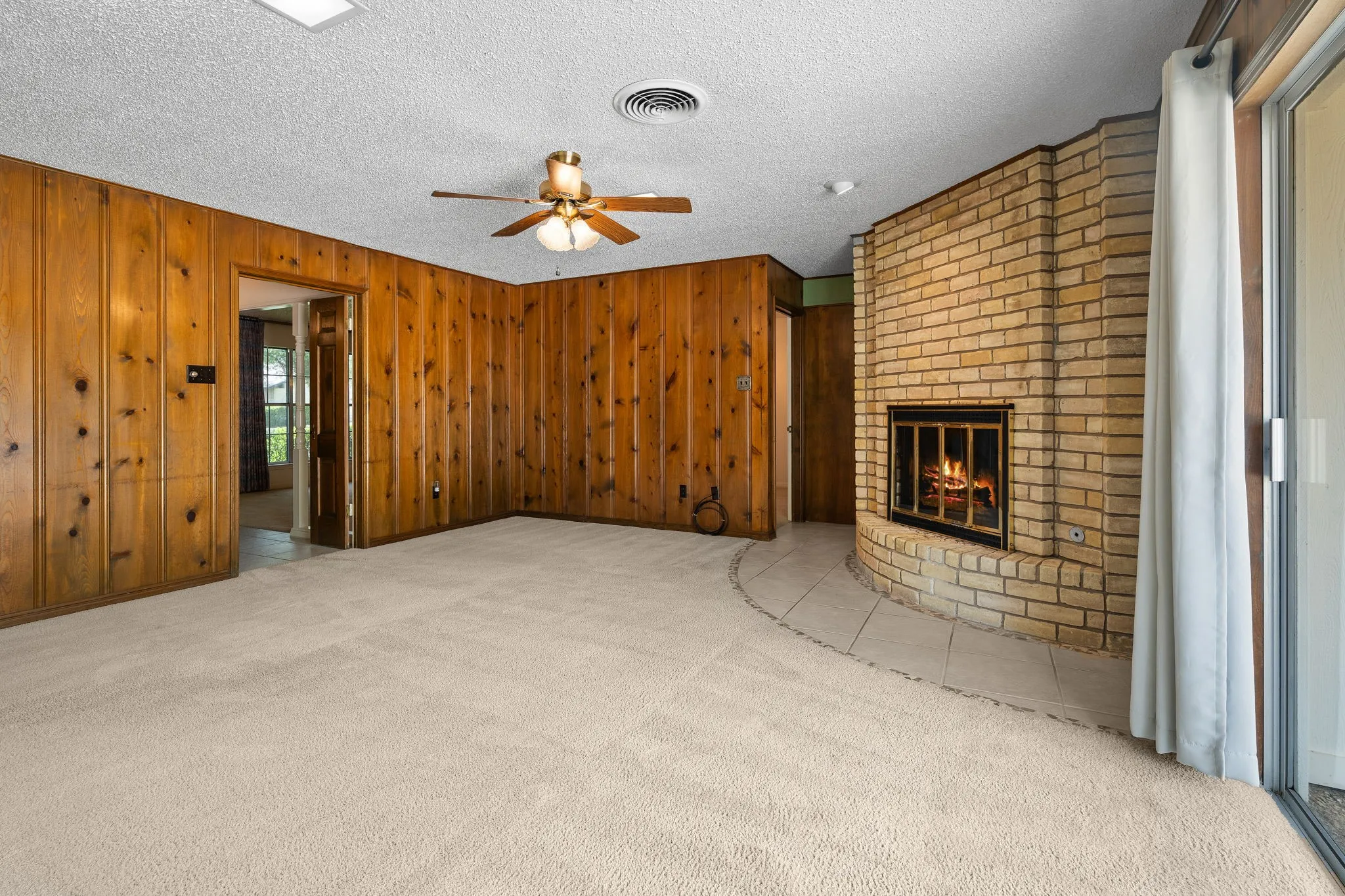 Corner brick fireplace with a black metal firebox, wood paneling, light-toned carpeting, and a ceiling fan with integrated lighting