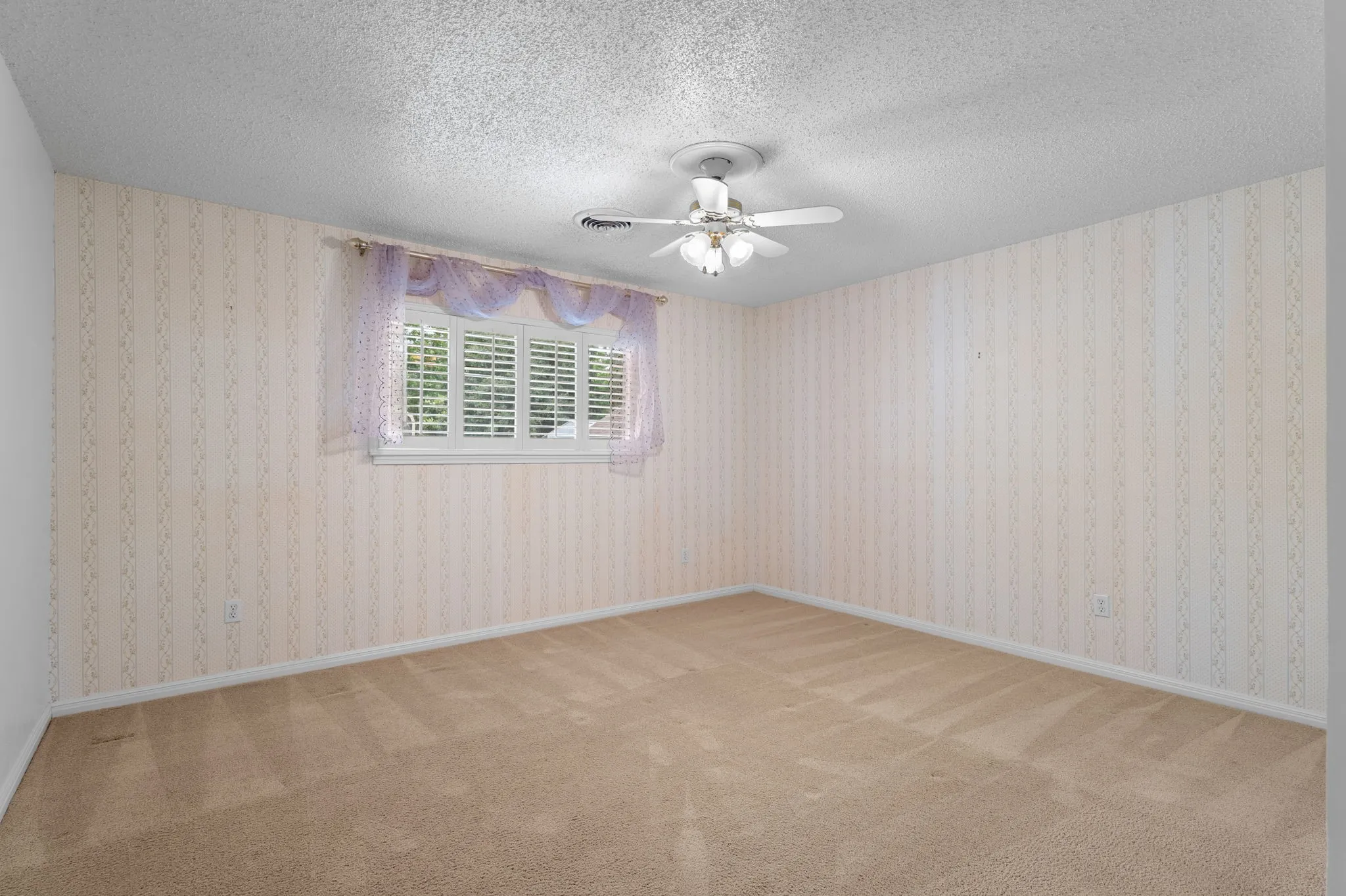 Room featuring textured carpeting, patterned wallpaper, a ceiling fan with integrated lighting, and a window with white louvered shutters