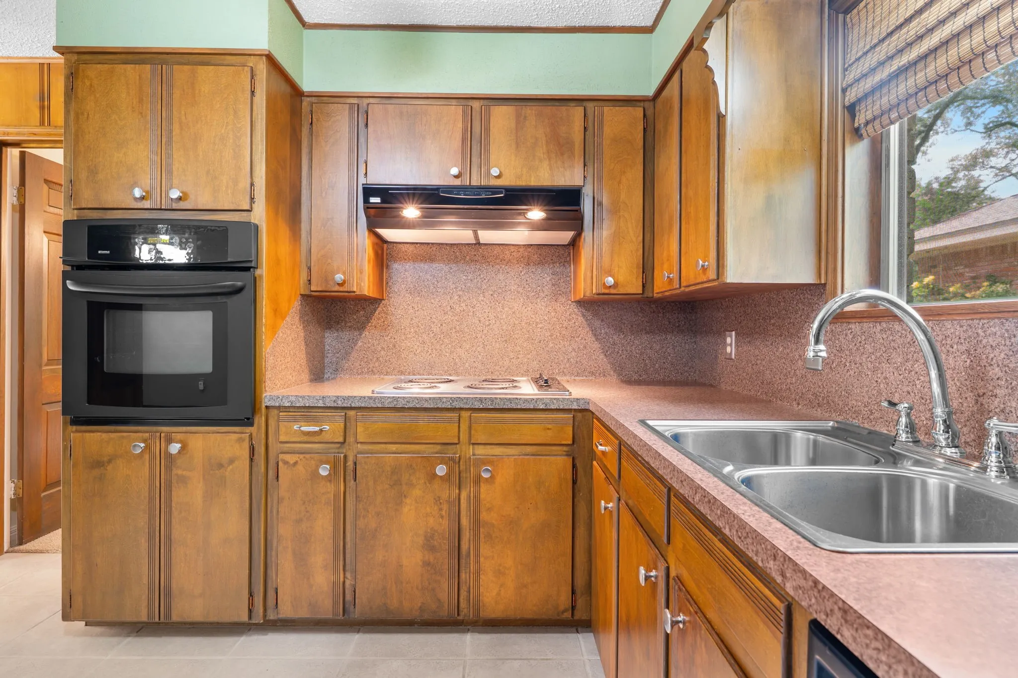 Kitchen featuring wood cabinetry, an integrated wall oven, a stainless steel double basin sink with a high-arc faucet, and light-colored tile flooring
