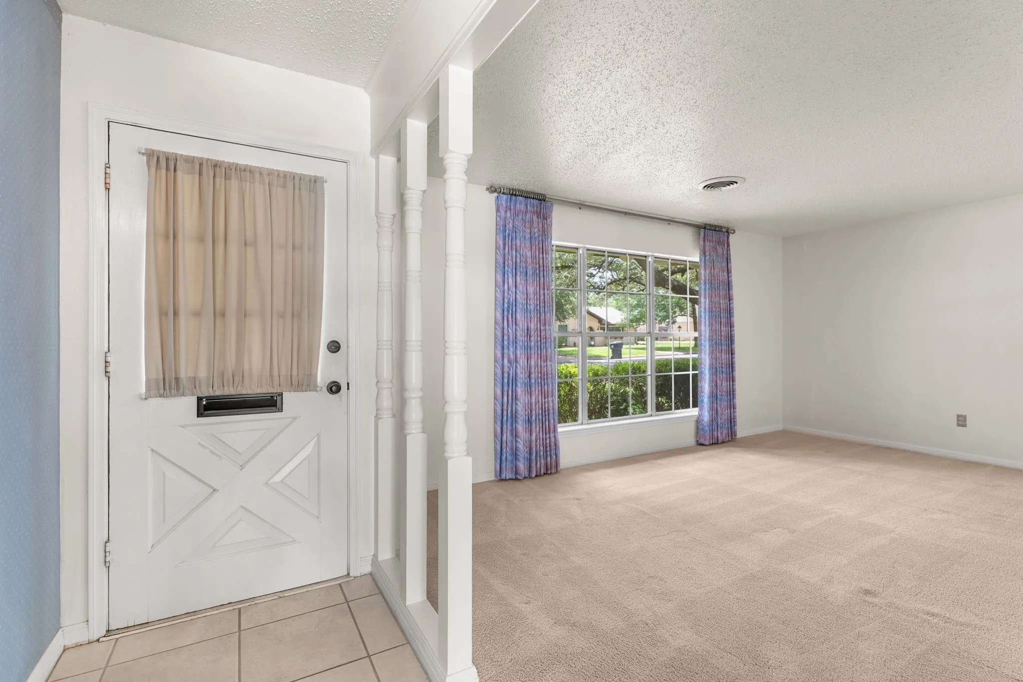 Inviting entryway featuring an ornate white front door and light-toned tile flooring, opening to a carpeted room with a large window and white walls