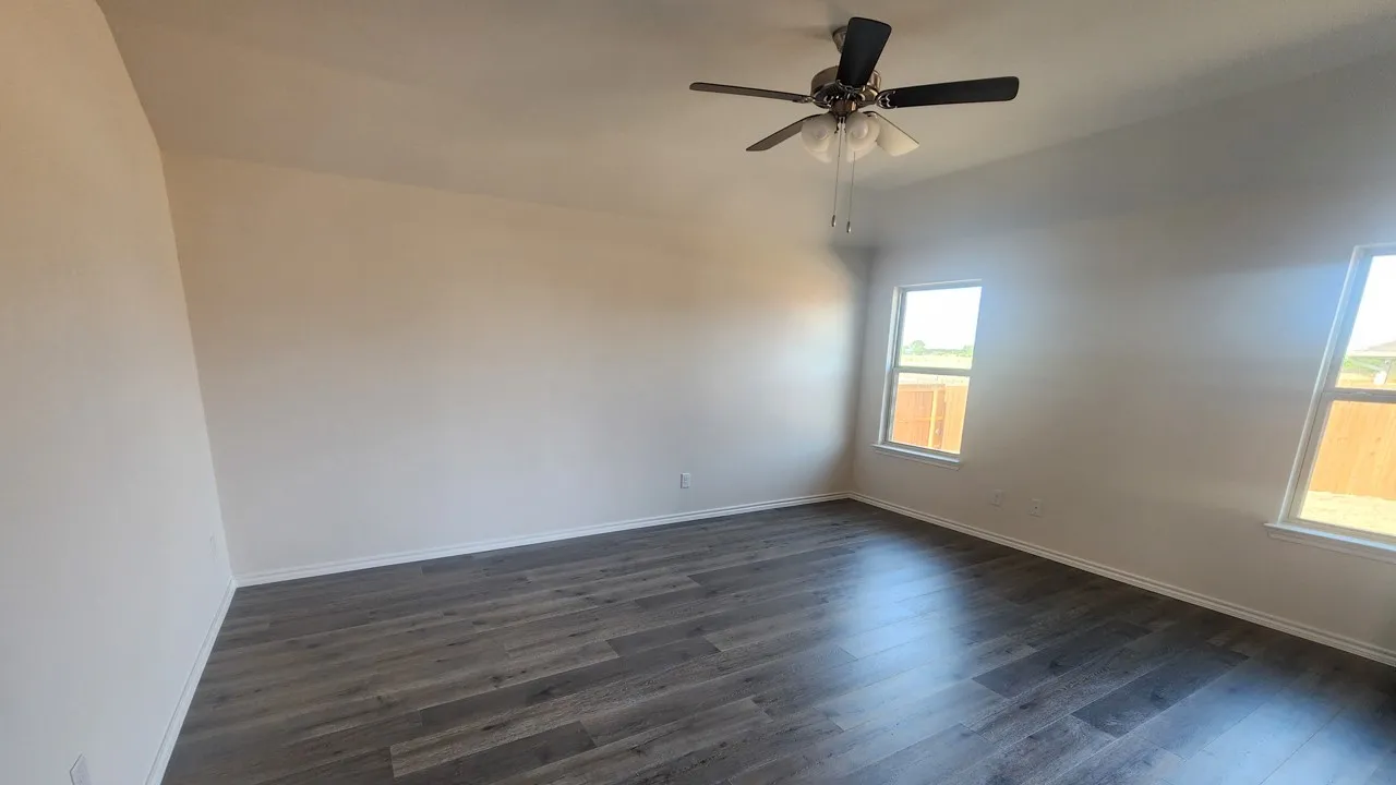 Spacious interior room featuring wood-finish flooring, neutral wall tones, multiple windows, a ceiling fan with integrated lighting, and white baseboards